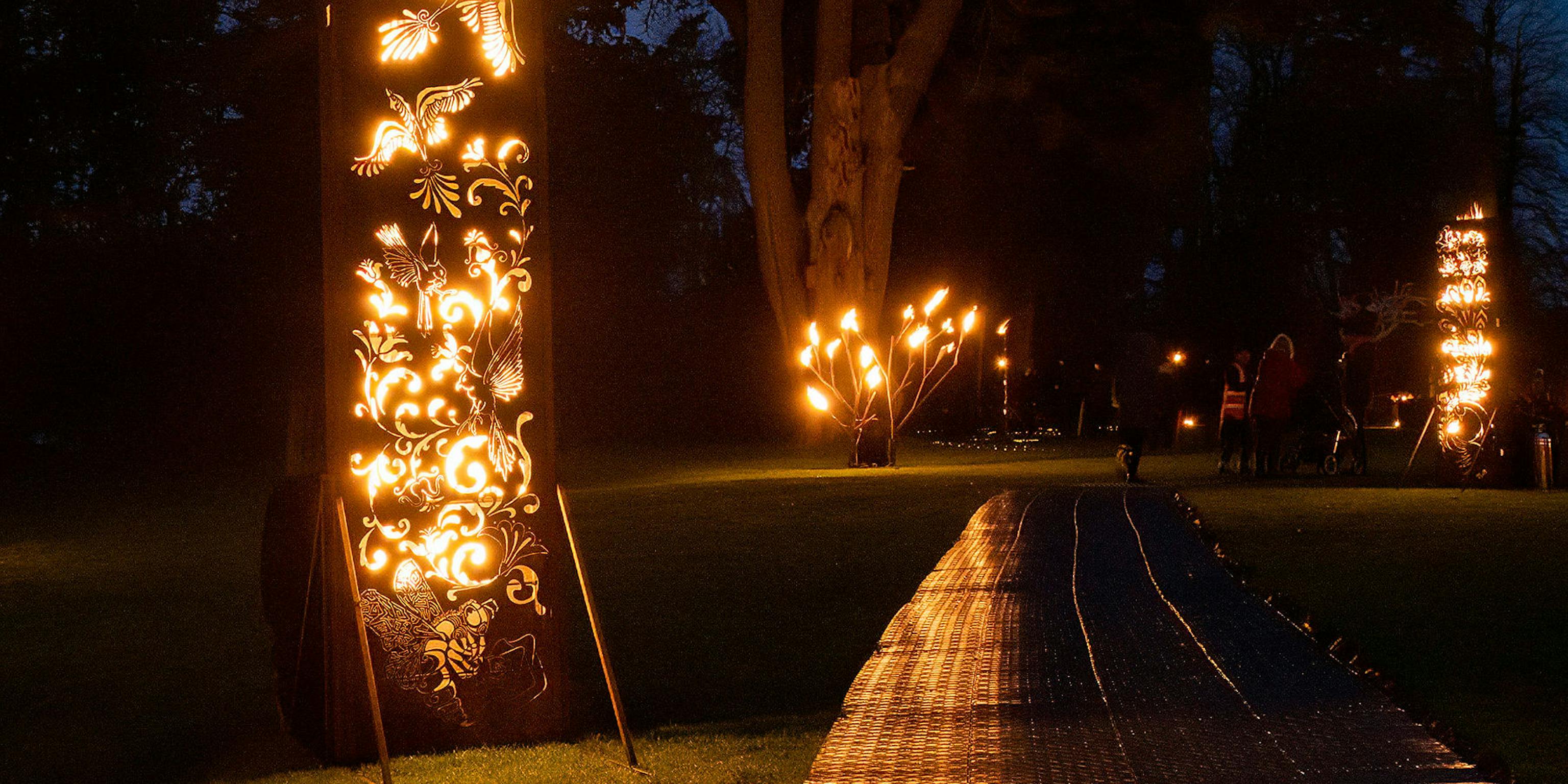 A pathway through a dark wood lit by fire sculptures along the side