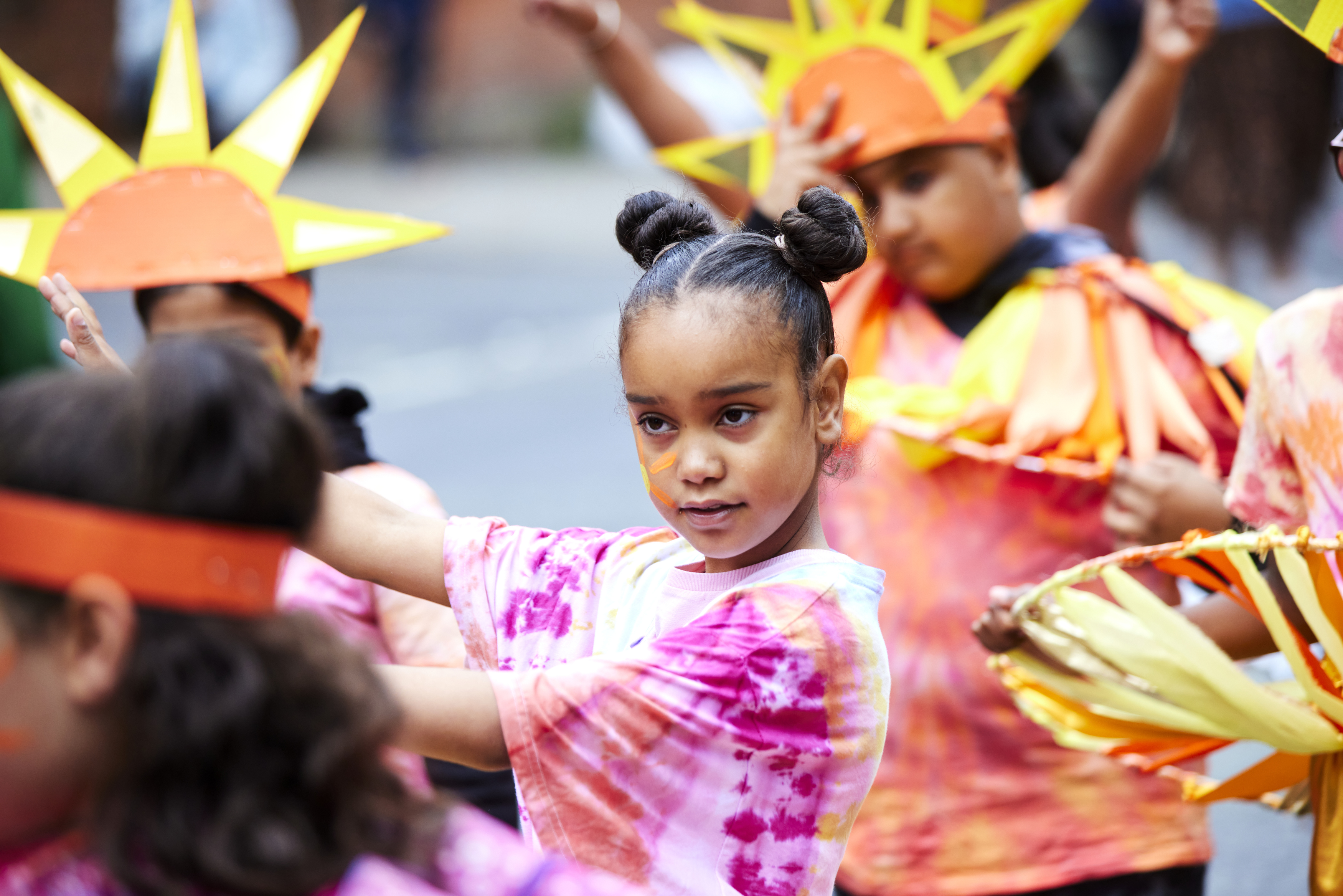 A young person with hair in two buns and a tie dye pink top dancing amongst other young people dress up as suns