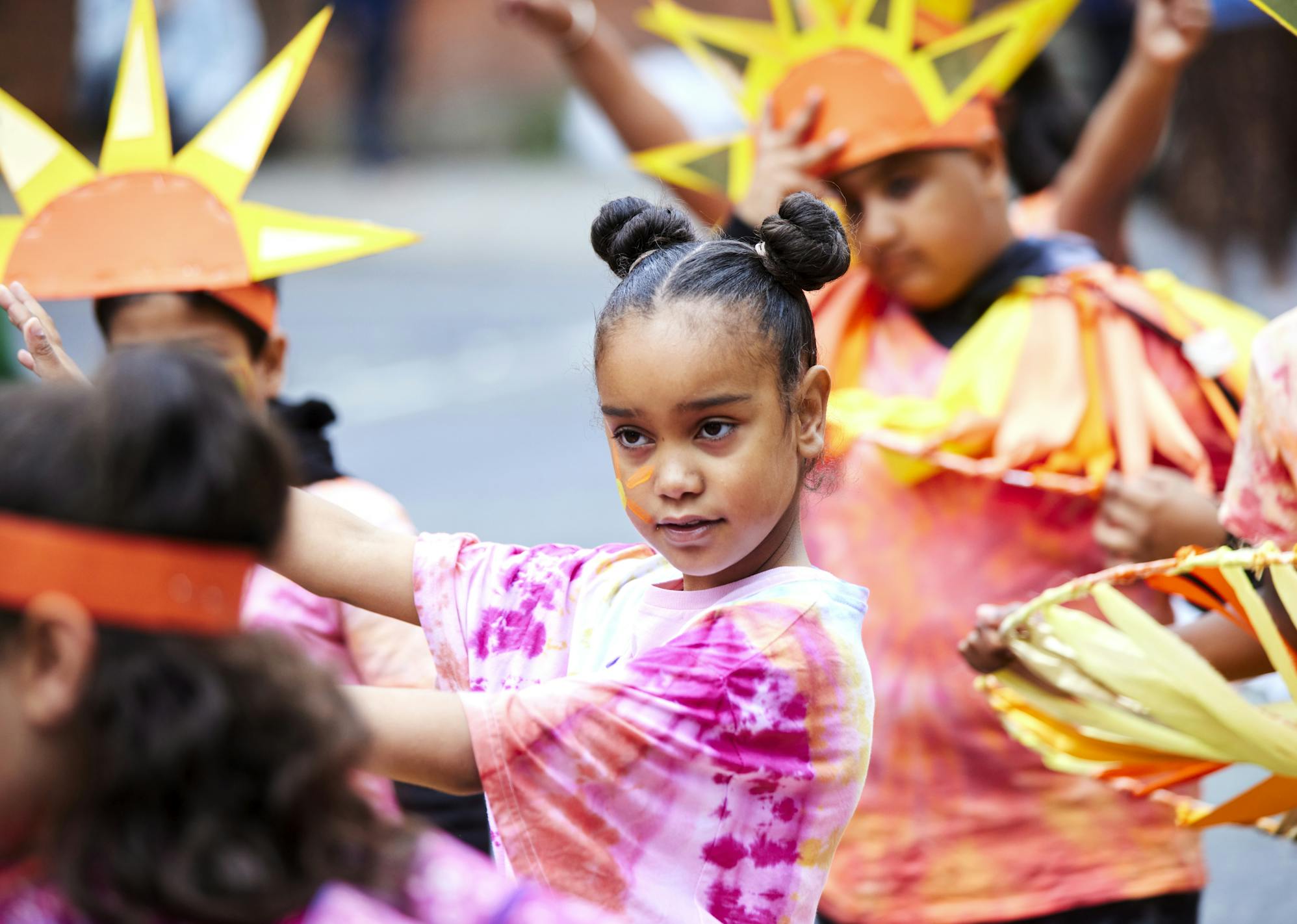 A young person with hair in two buns and a tie dye pink top dancing amongst other young people dress up as suns