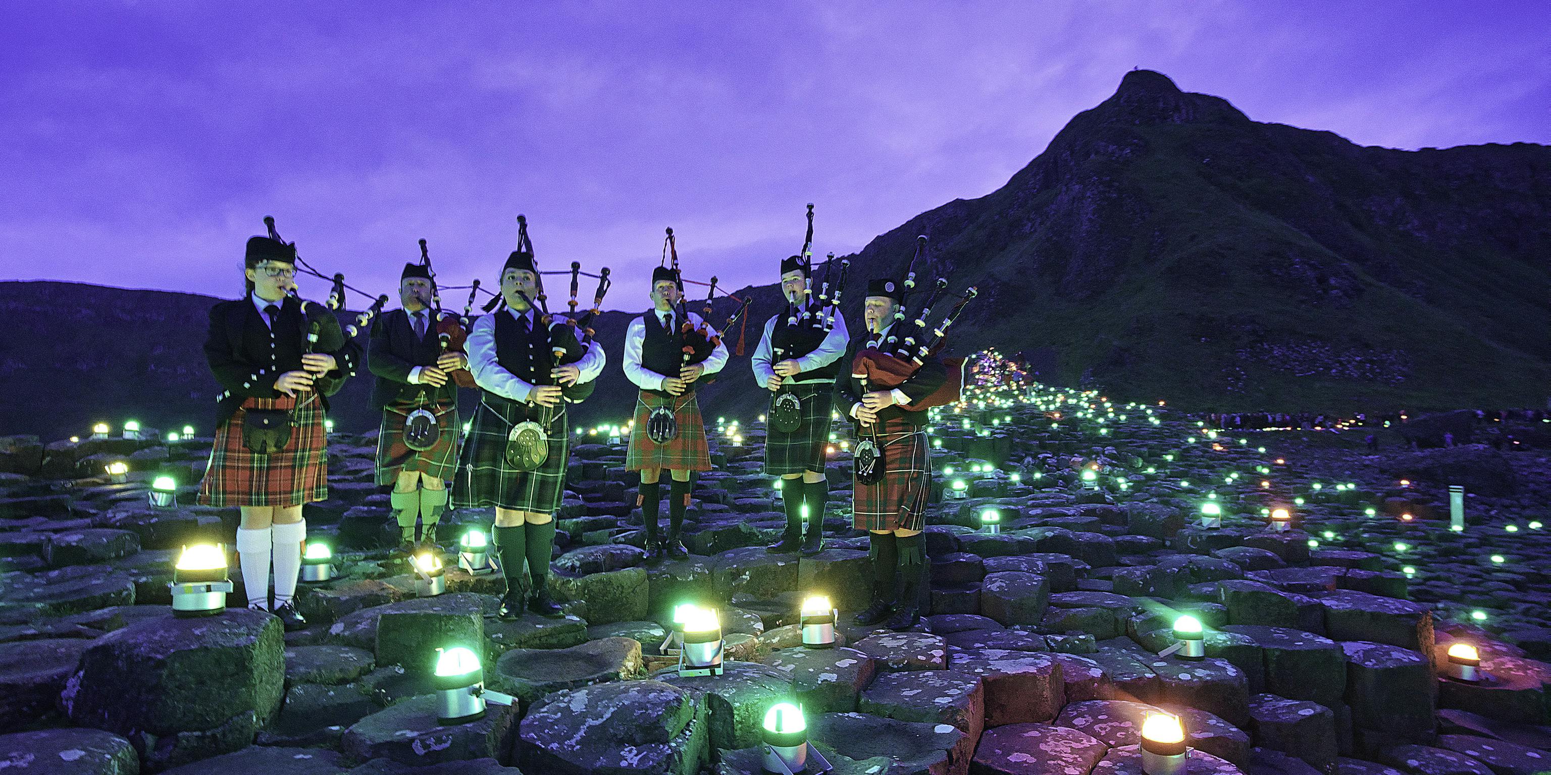 A group of six bagpipe players standing on the stones of Giants Causeway surrounded by different coloured geolights