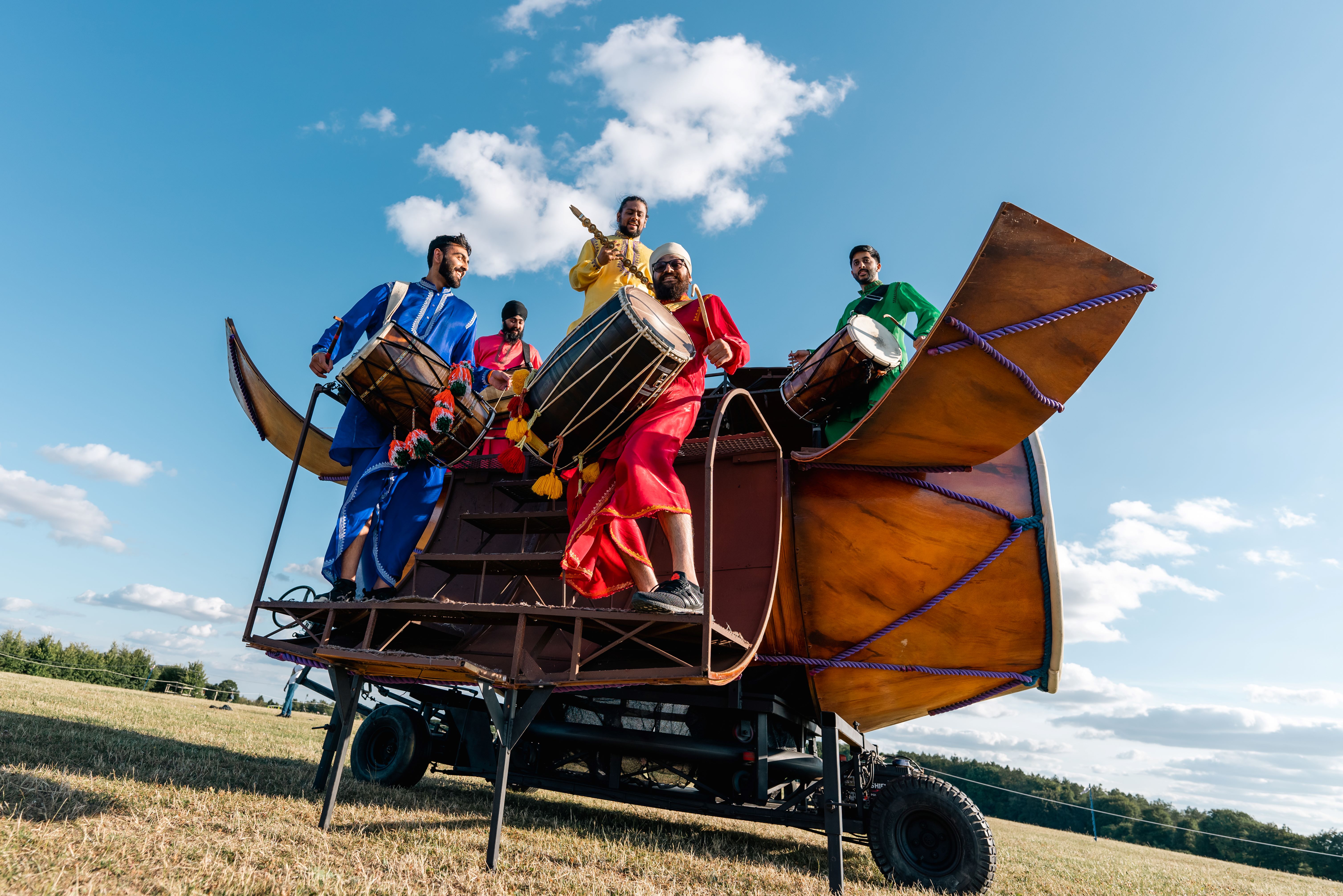 Six drummers in brightly coloured clothing standing inside a giant Dhol drum
