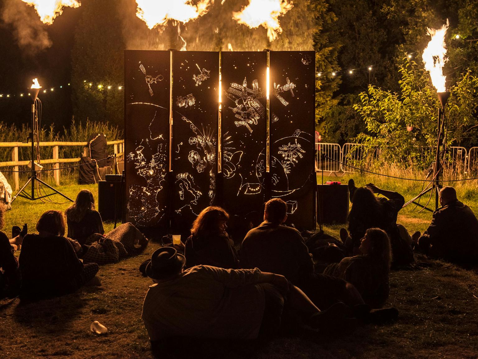 A group of people sitting on the grass watching a space-themed fir sculpture at night