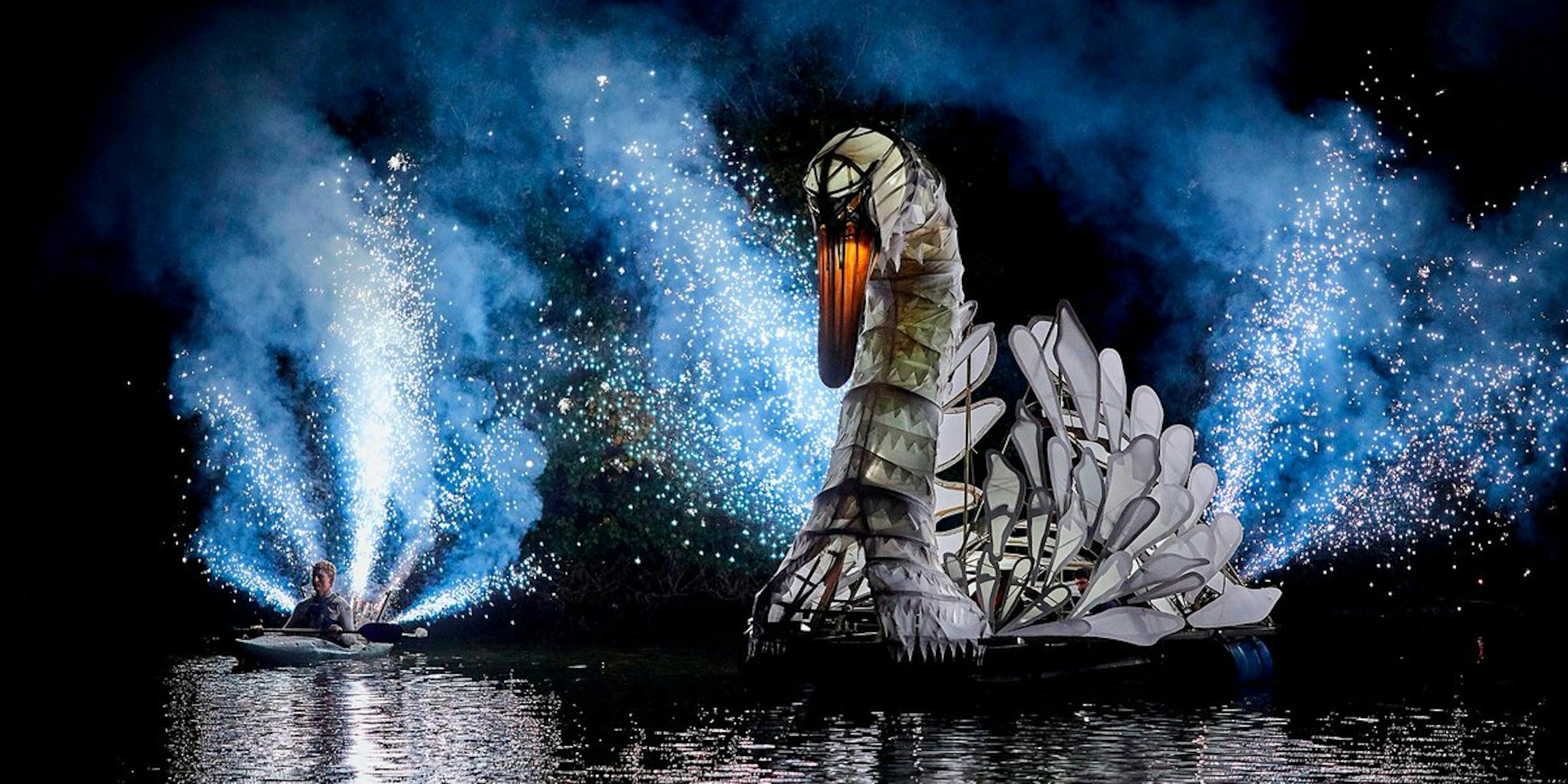 A structure of lit-up swan in water at night surrounded by pyro and a person in a canoe