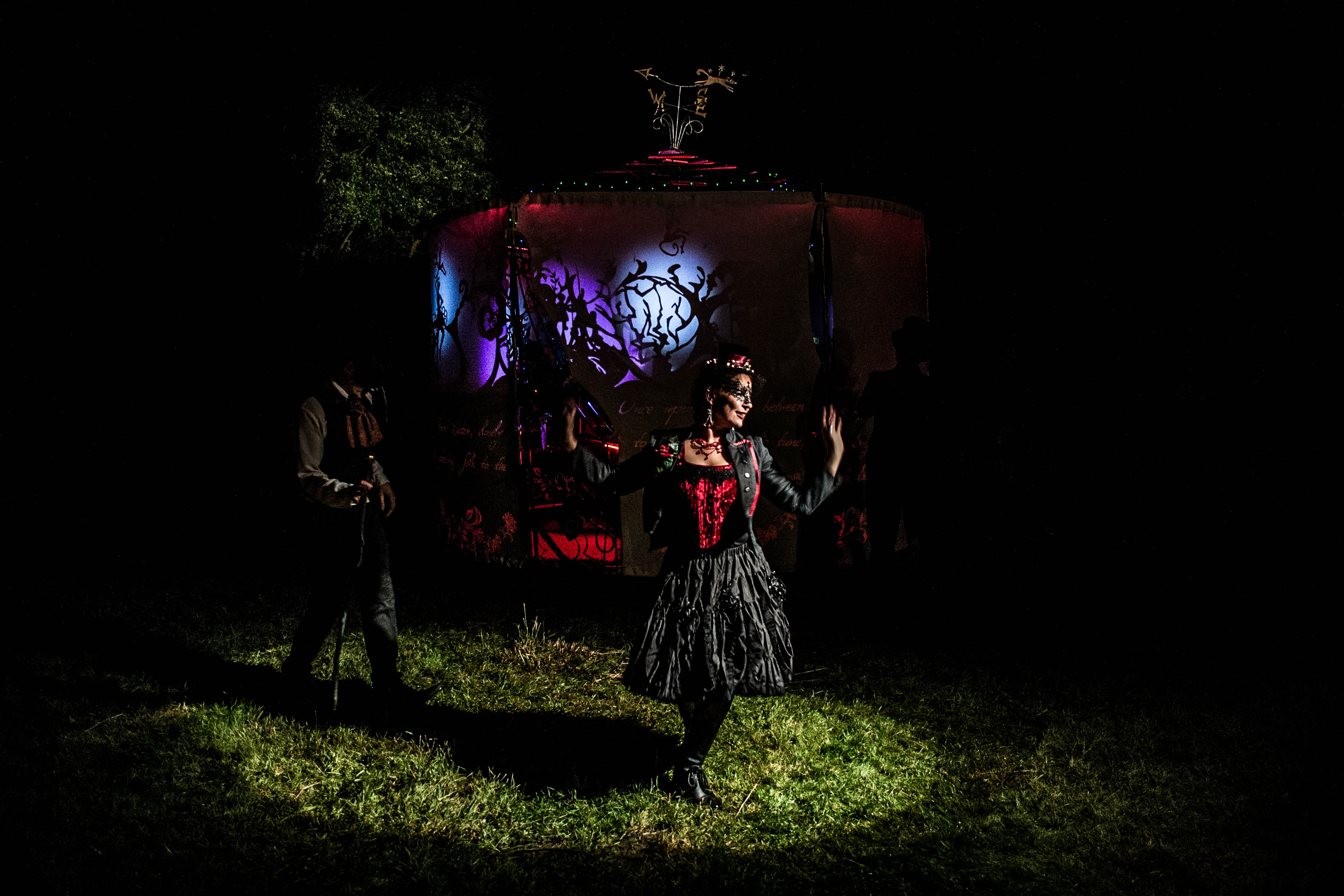 A woman in a black dress and mask standing in a spotlight on grass in front of a blue light with shadow puppets on it