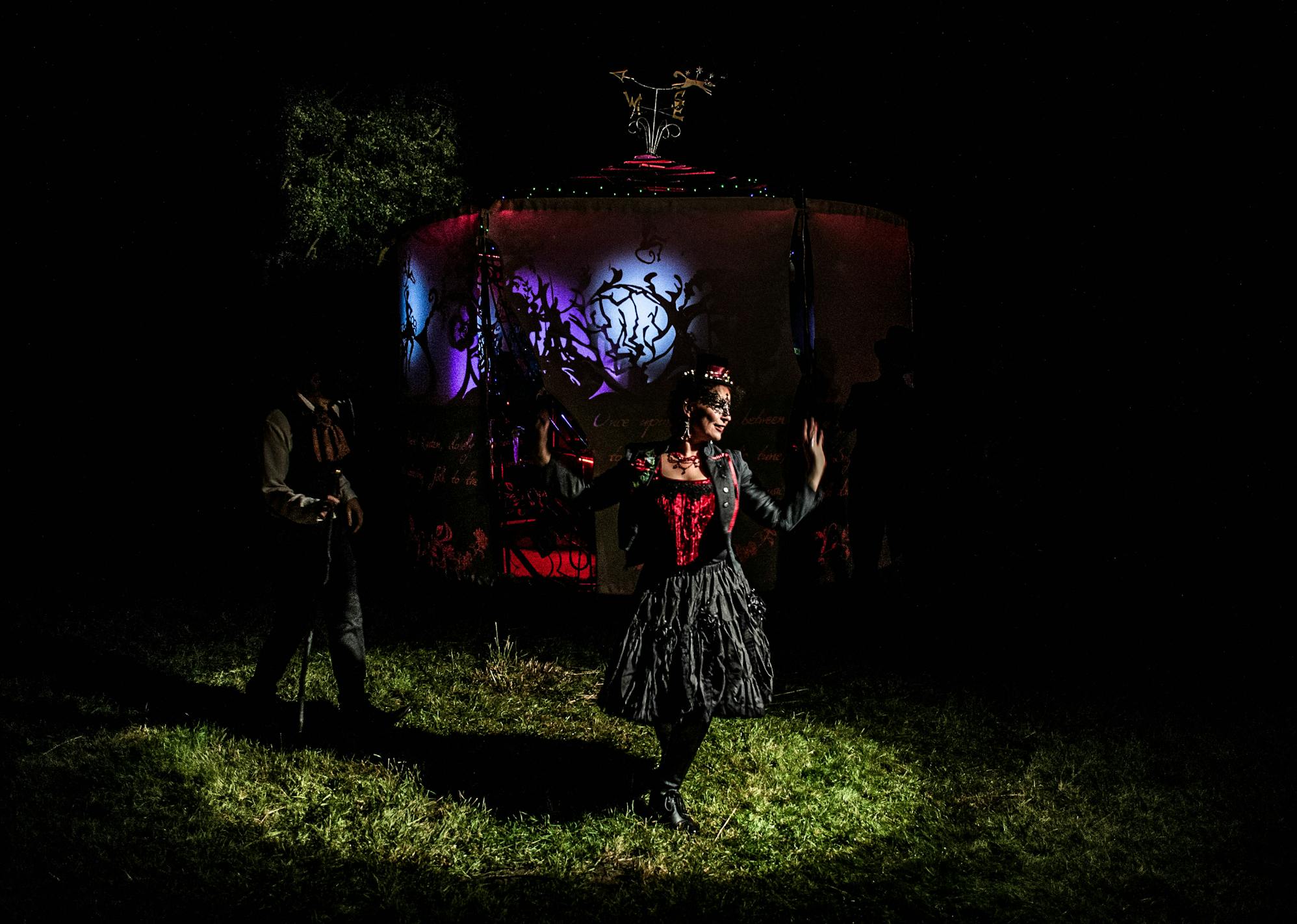 A woman in a black dress and mask standing in a spotlight on grass in front of a blue light with shadow puppets on it