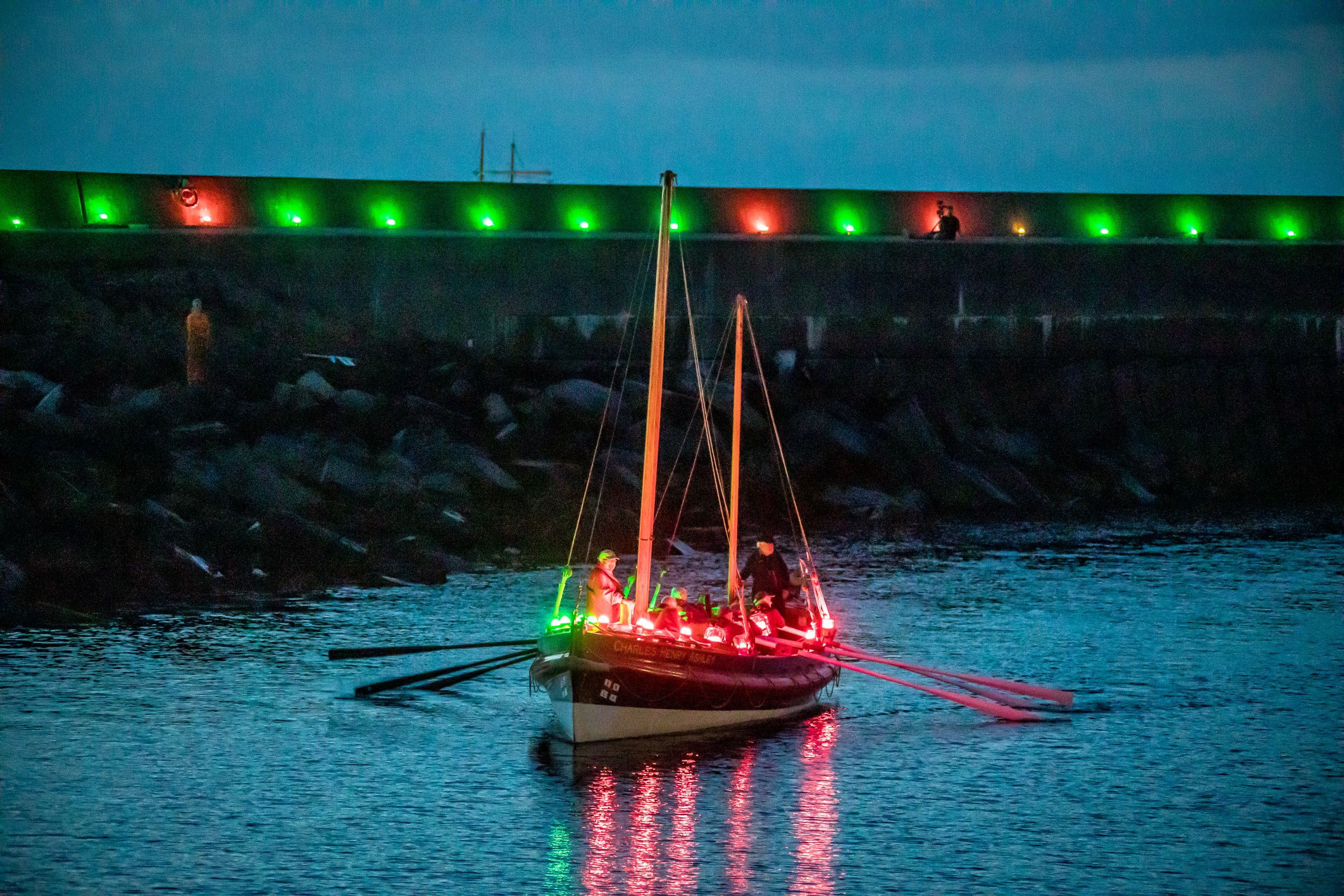 A boat filled with Red and Green Geolights leaving a harbour that is also lined in Geolights