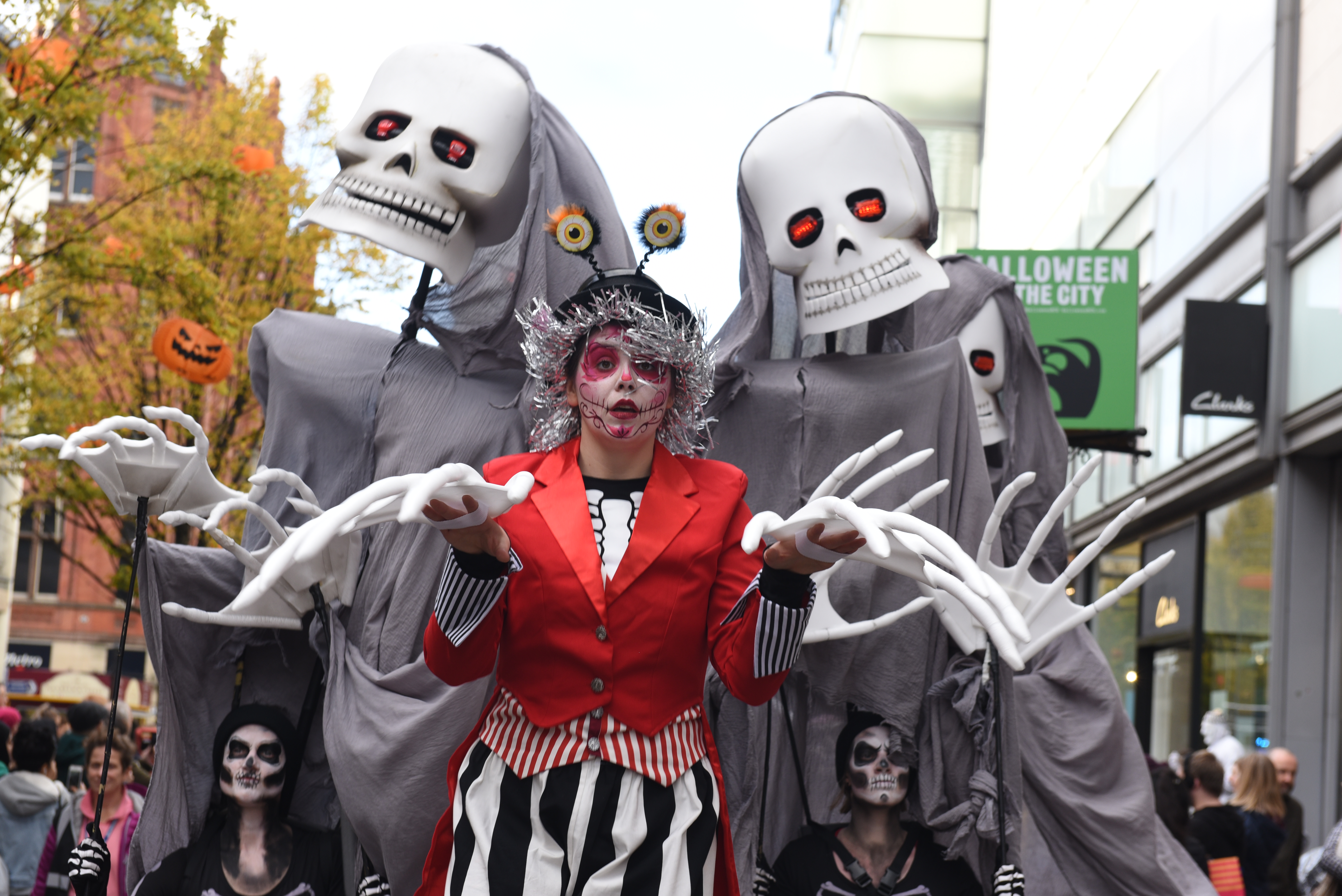 A performer in a red jacket and black and white striped trousers leads two large skeleton puppets through a town centre