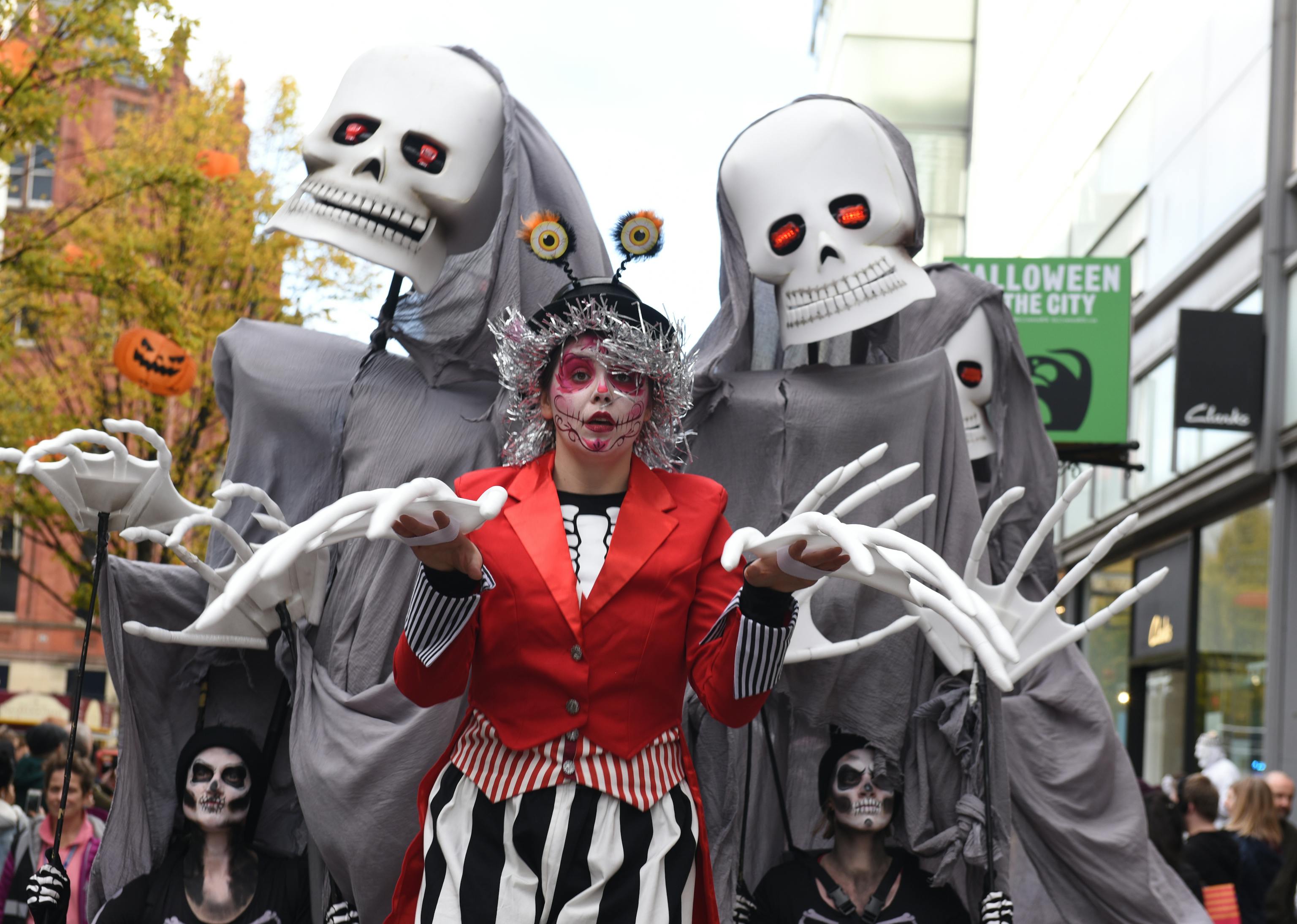 A performer in a red jacket and black and white striped trousers leads two large skeleton puppets through a town centre
