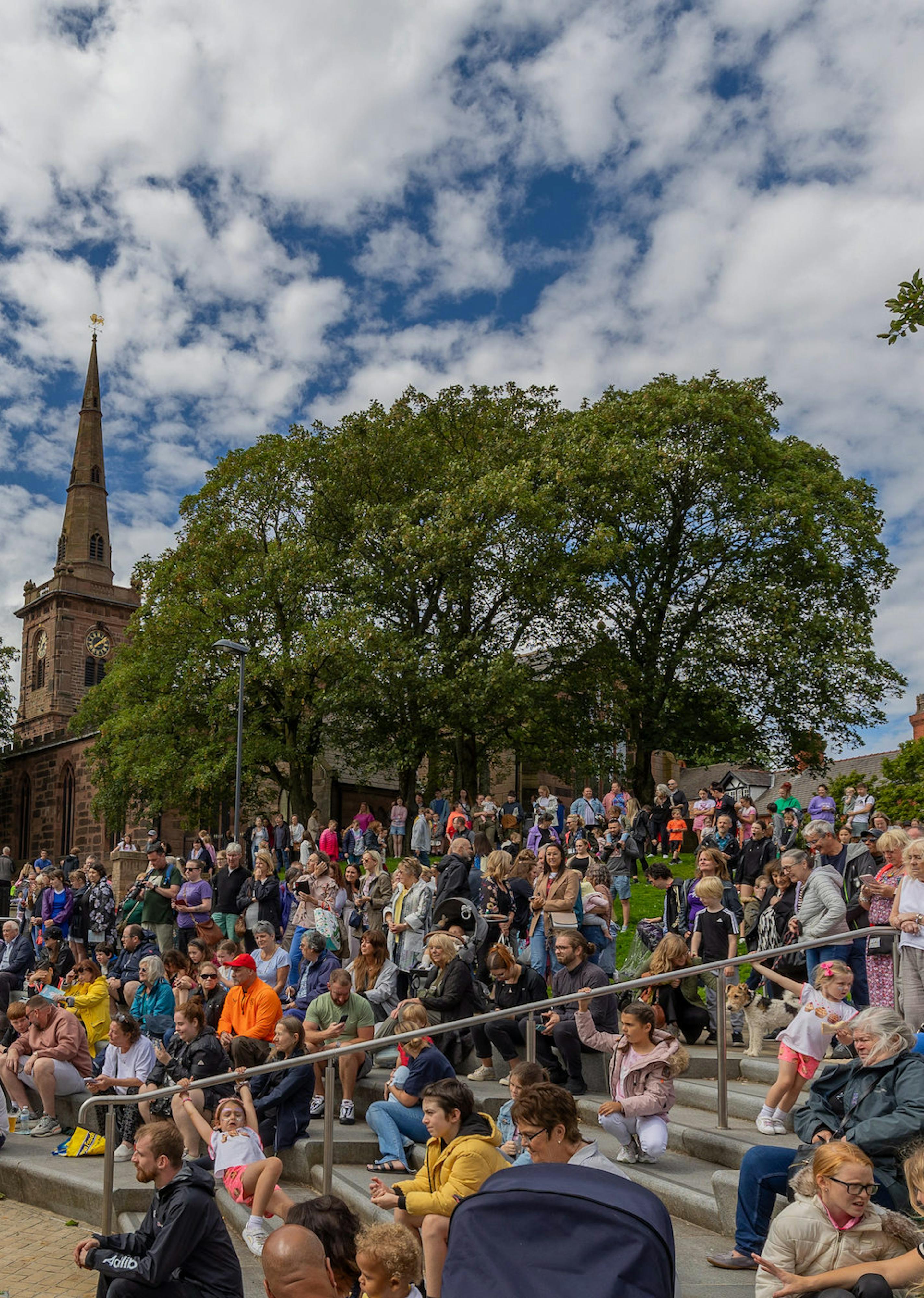 A crowd of people sitting on steps watching a performance with cloudy skies over head