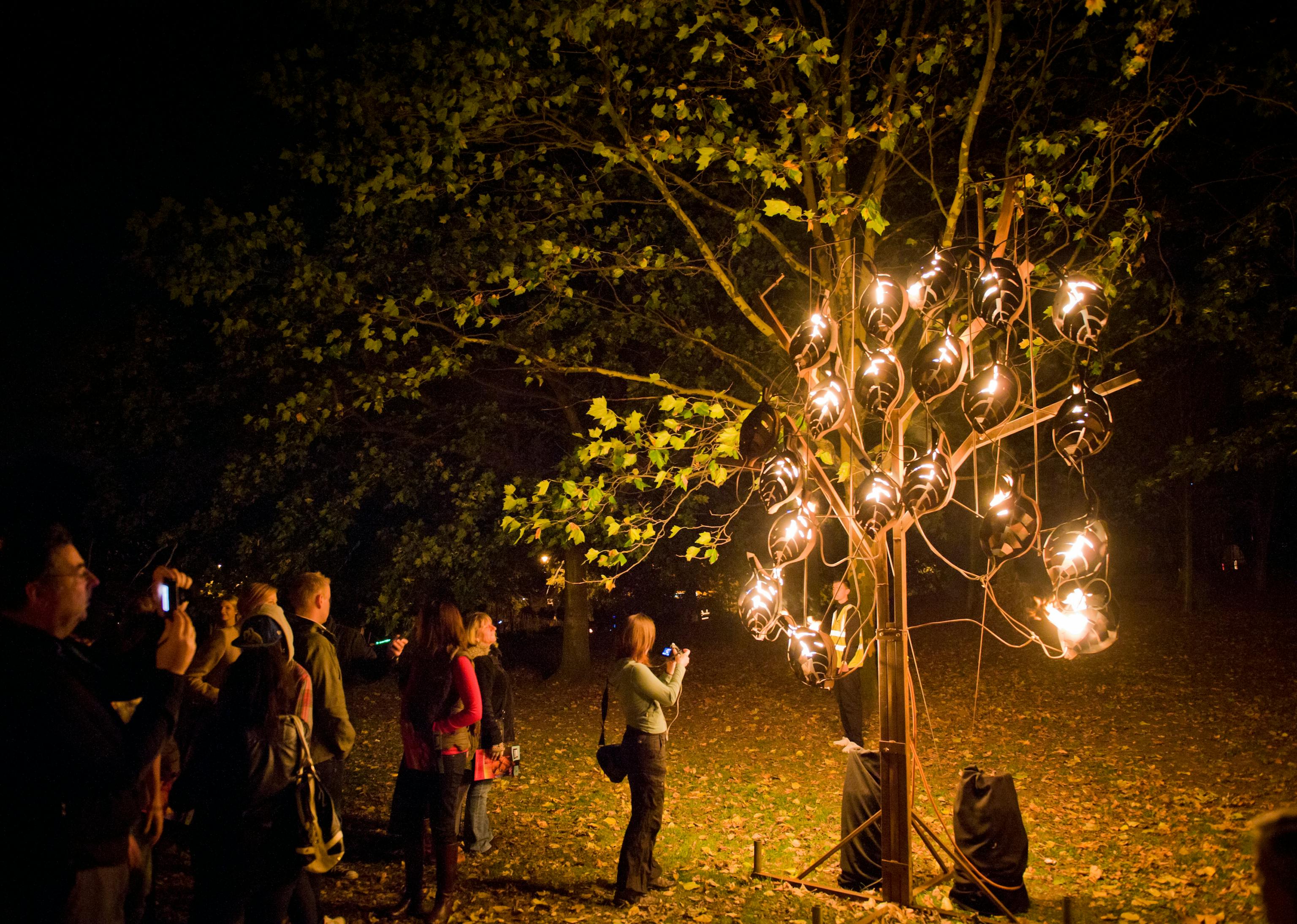 A group of people gathered around a fire tree sculpture in a woodland area, taking photographs