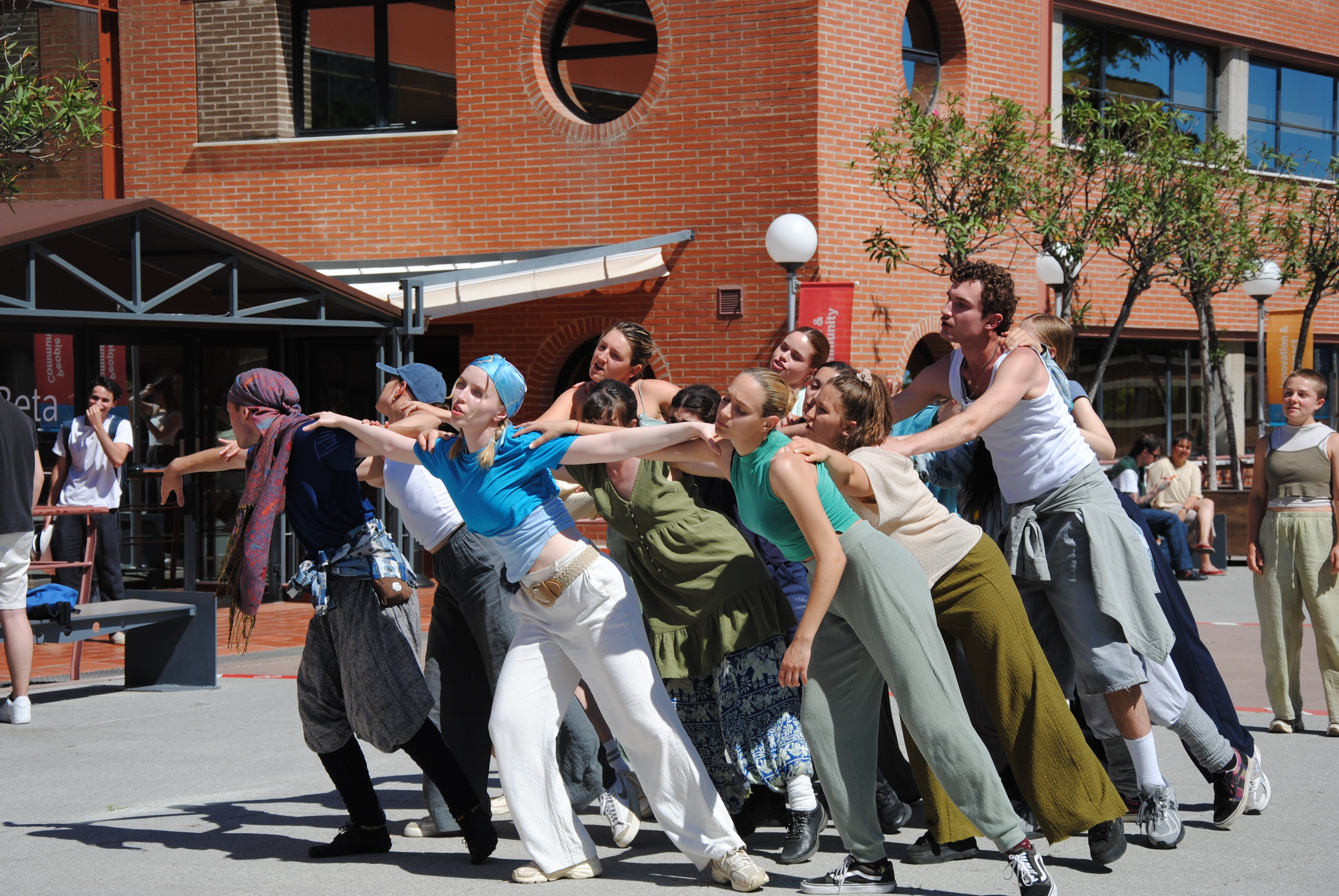 Group of dancers performing a synchronised lean outdoors in front of a brick building on a sunny day.