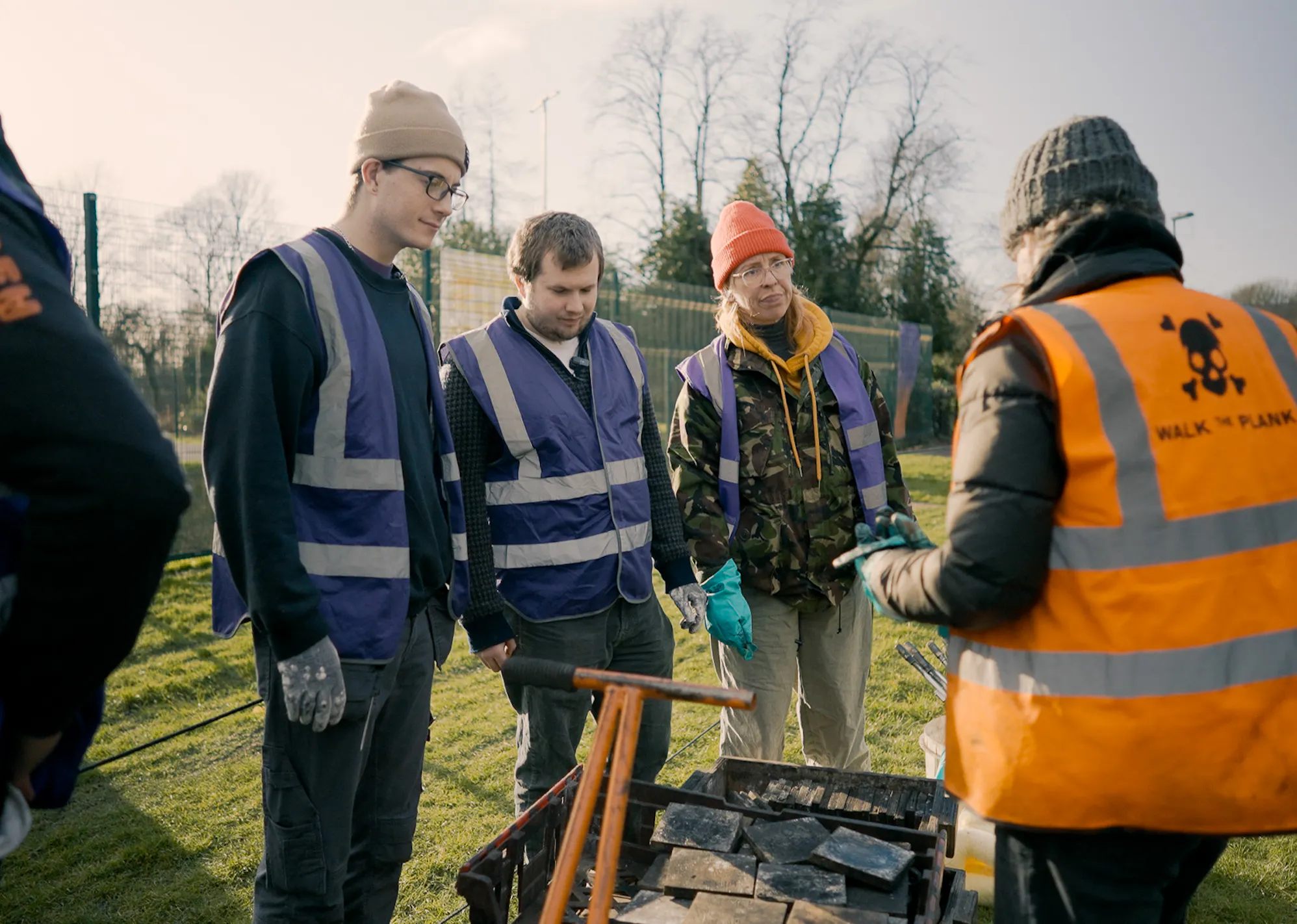 Three people in purple high-vis jackets listening to someone in a orange high-vis jacket outdoors.