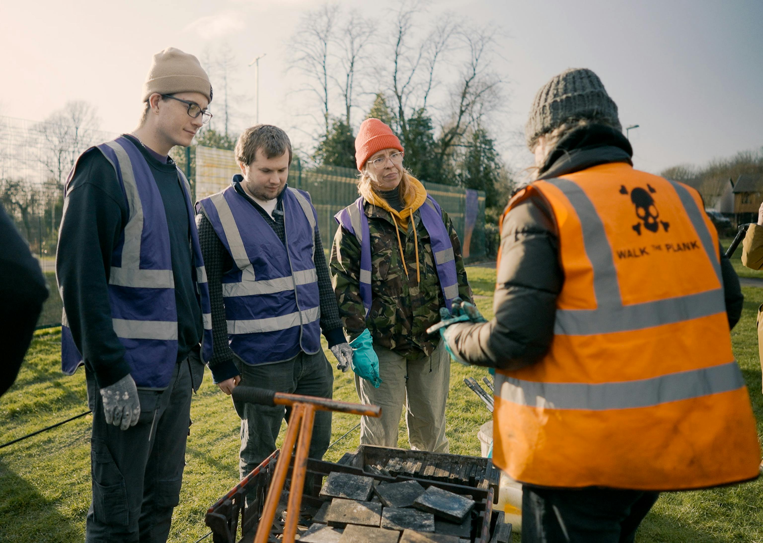 Three people in purple high-vis jackets listening to someone in a orange high-vis jacket outdoors.