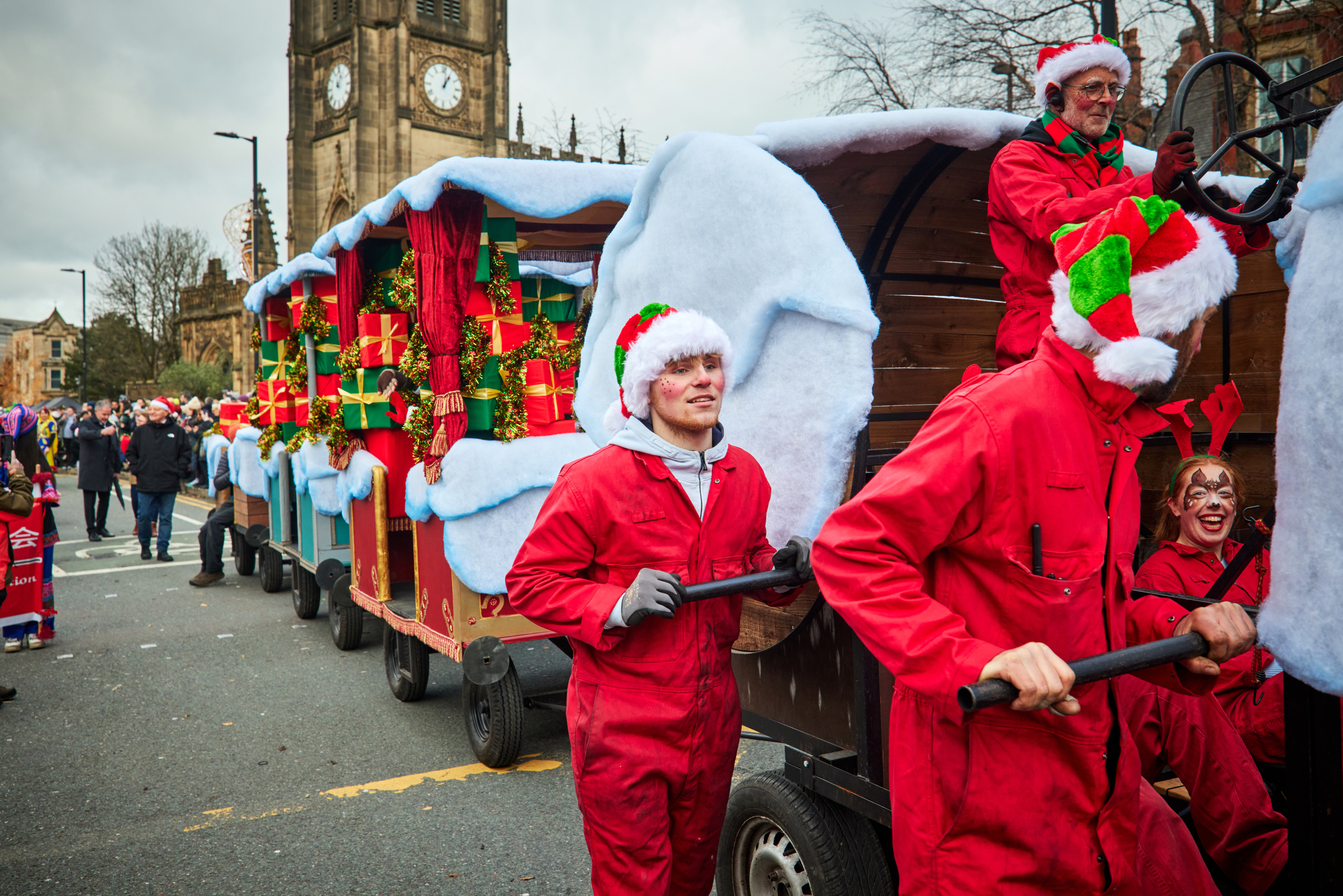 A peddle powered three carriage procession train covered in fabric snow, decorative presents and people in red jumpsuits and Santa's hats. 
