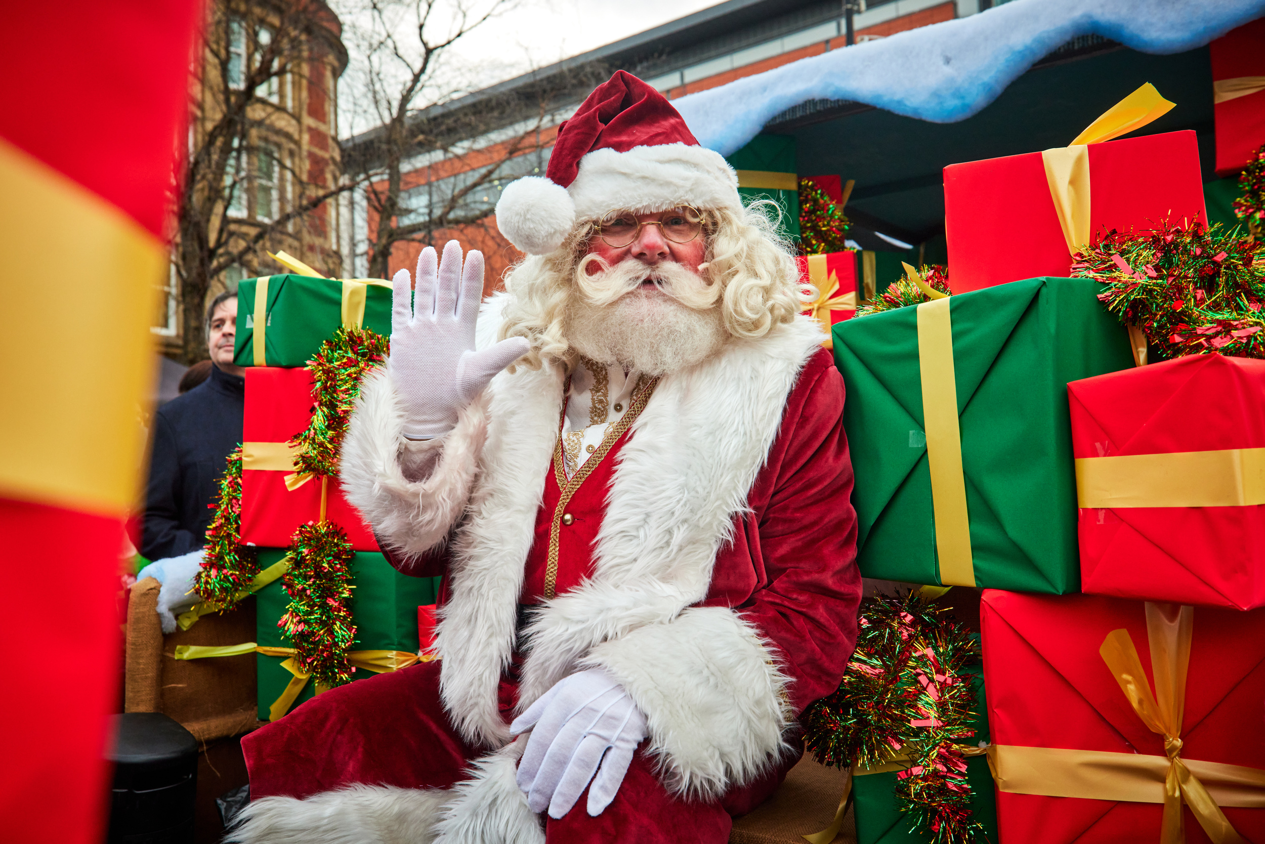 A performer dressed as Santa Clause with a white beard, a red and white outfit and hat, surrounded by presents. 