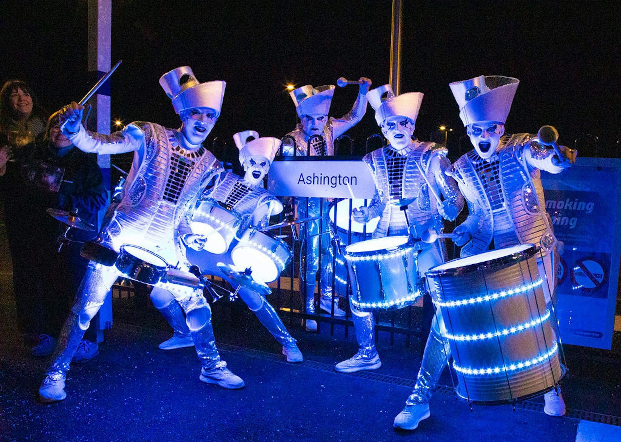 A Group of Drummers dressed all in white posing by a sign that reads 'Ashington'