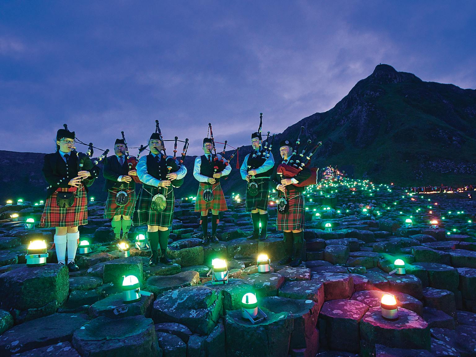 A group of six bagpipe players standing on the stones of Giants Causeway surrounded by different coloured geolights