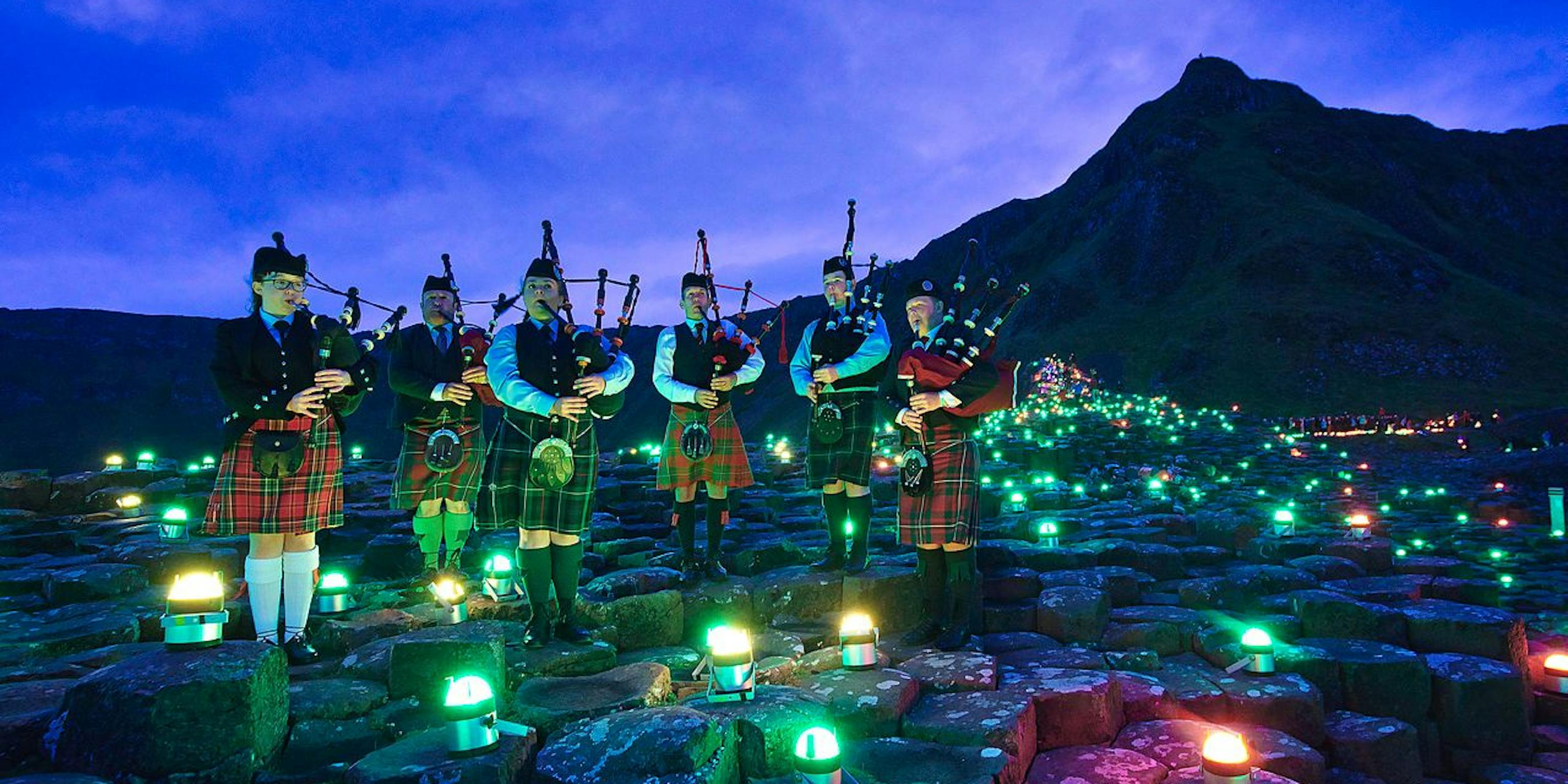 A group of bagpipe players wearing kilts standing on the stones of Giants Causeway surrounded by coloured lights