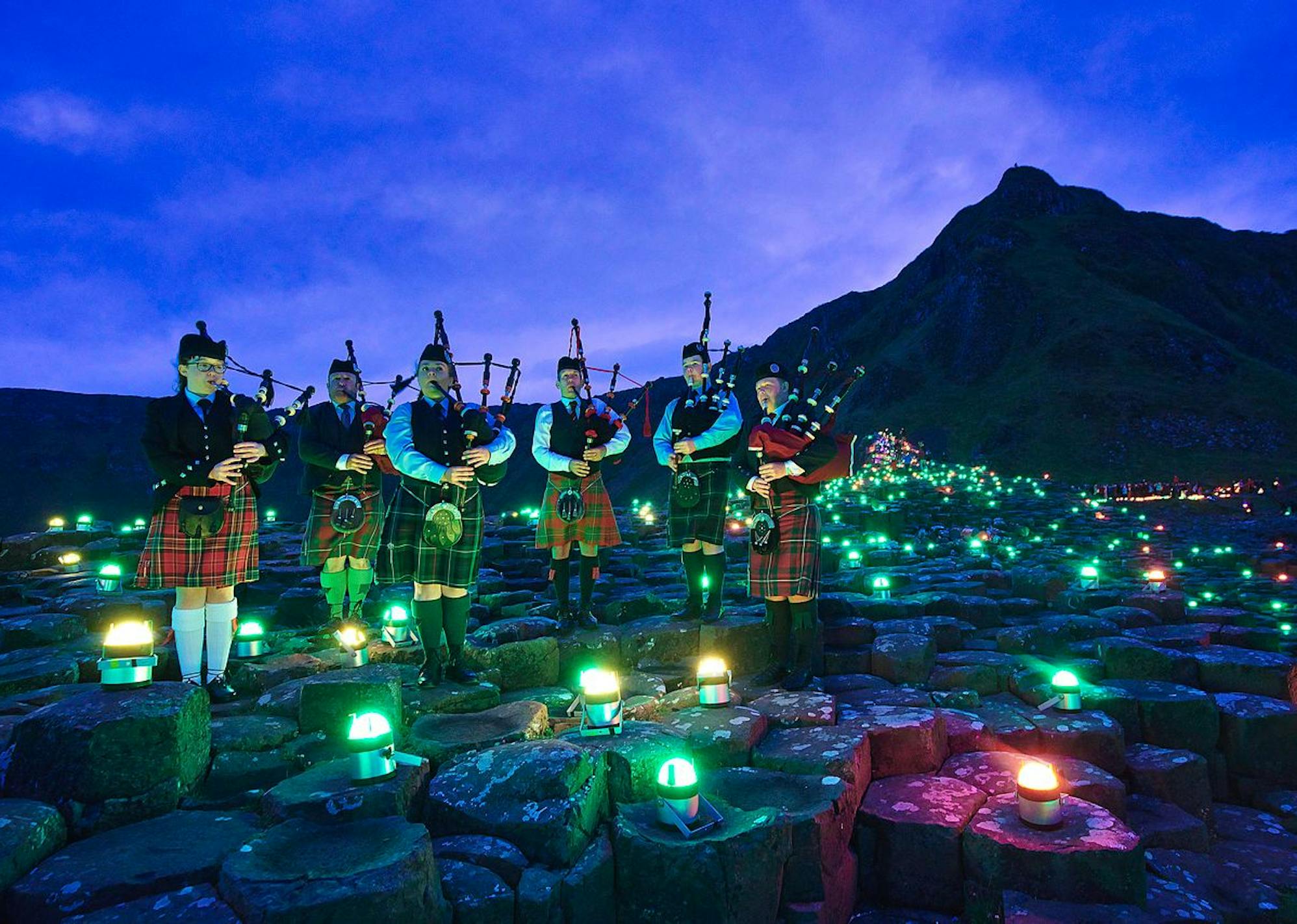 A group of bagpipe players wearing kilts standing on the stones of Giants Causeway surrounded by coloured lights