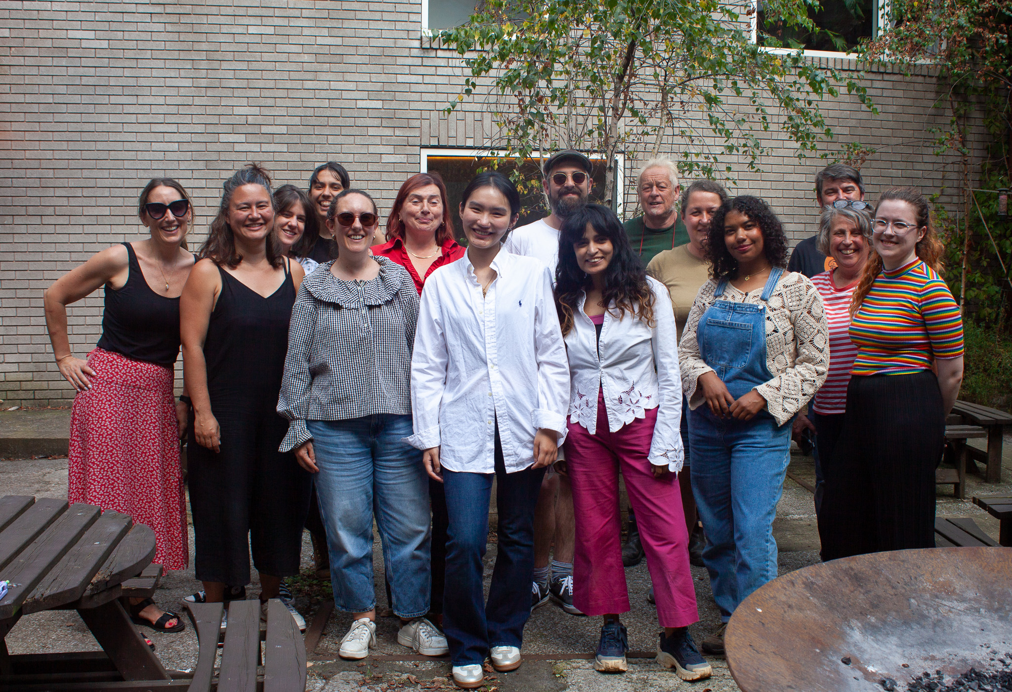 A work of work colleagues all posing in a group for a photo in a yard with wooden tables and chairs in the background. 