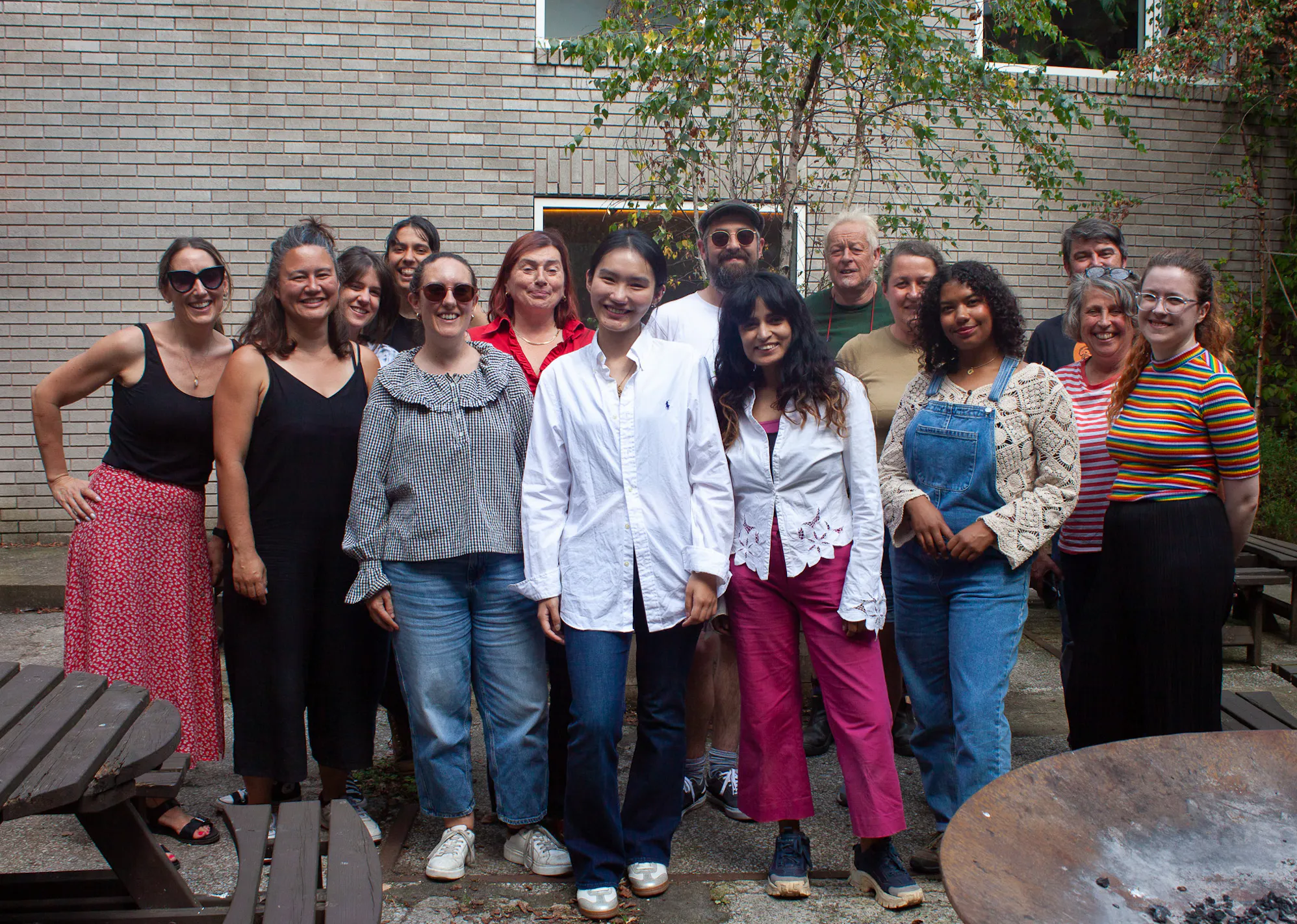A work of work colleagues all posing in a group for a photo in a yard with wooden tables and chairs in the background.