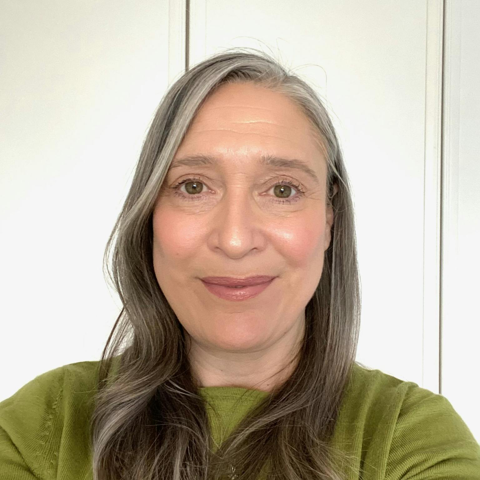 A headshot of a person, she is wearing a green top and standing in front of a white wall