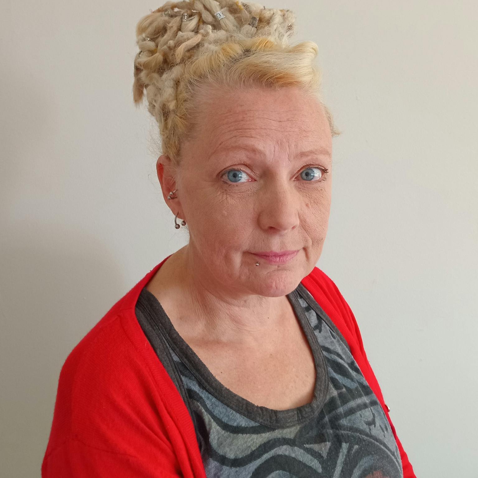 A headshot of a person in front of a white background. She has blonde hair in a bun and is wearing a red cardigan over a patterned grey top.
