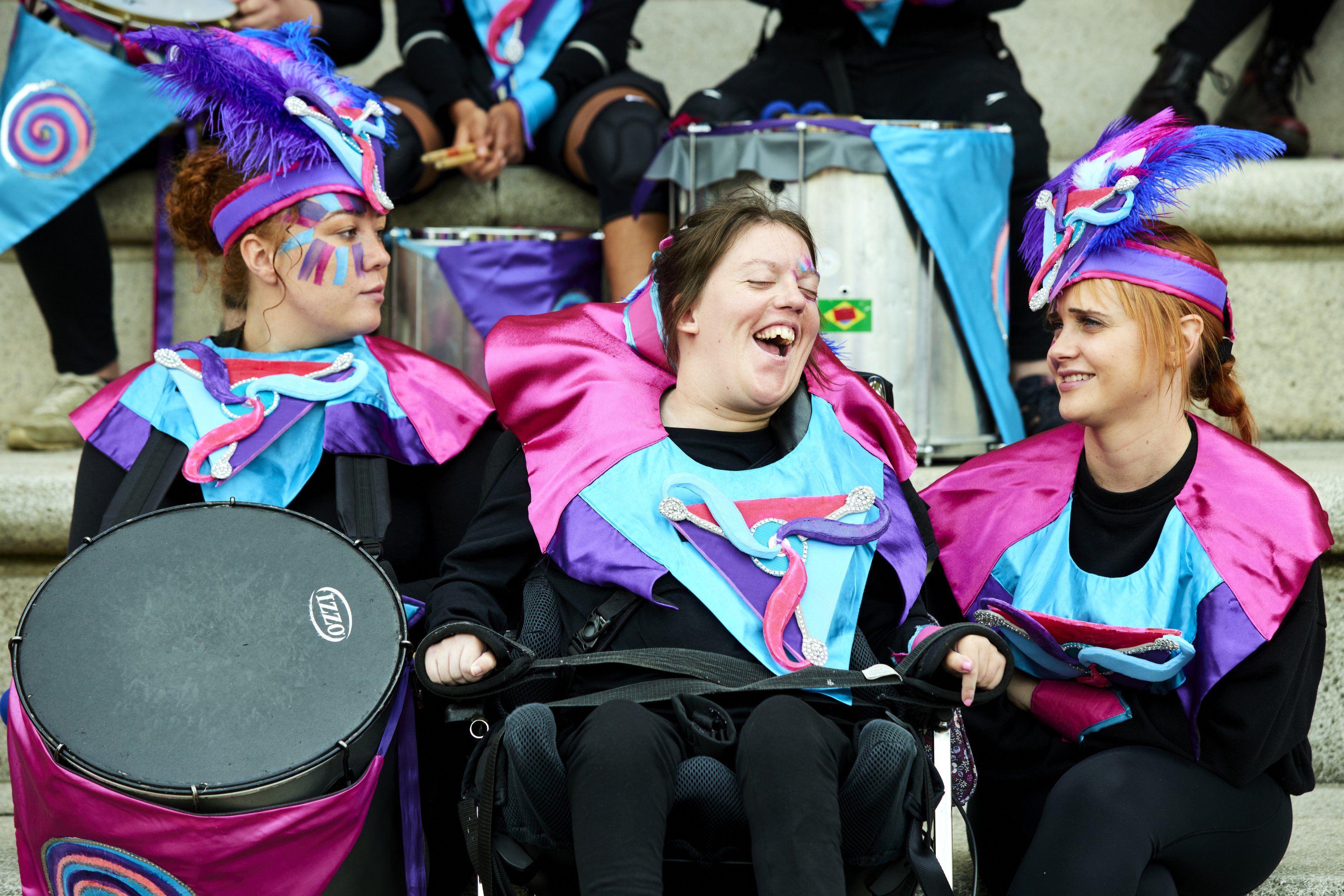 Three performers wearing a pink, purple and blue outfit and holding drums