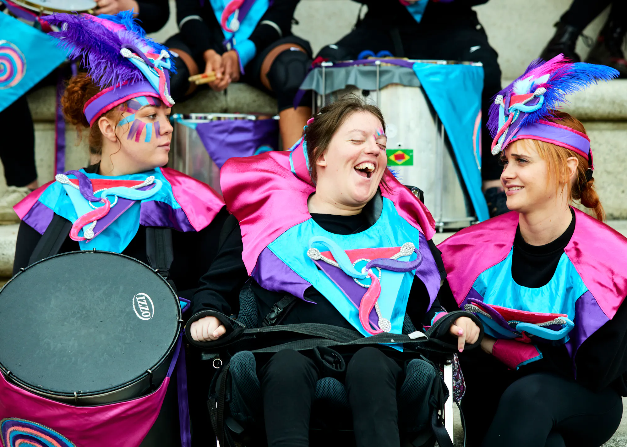 Three performers wearing a pink, purple and blue outfit and holding drums