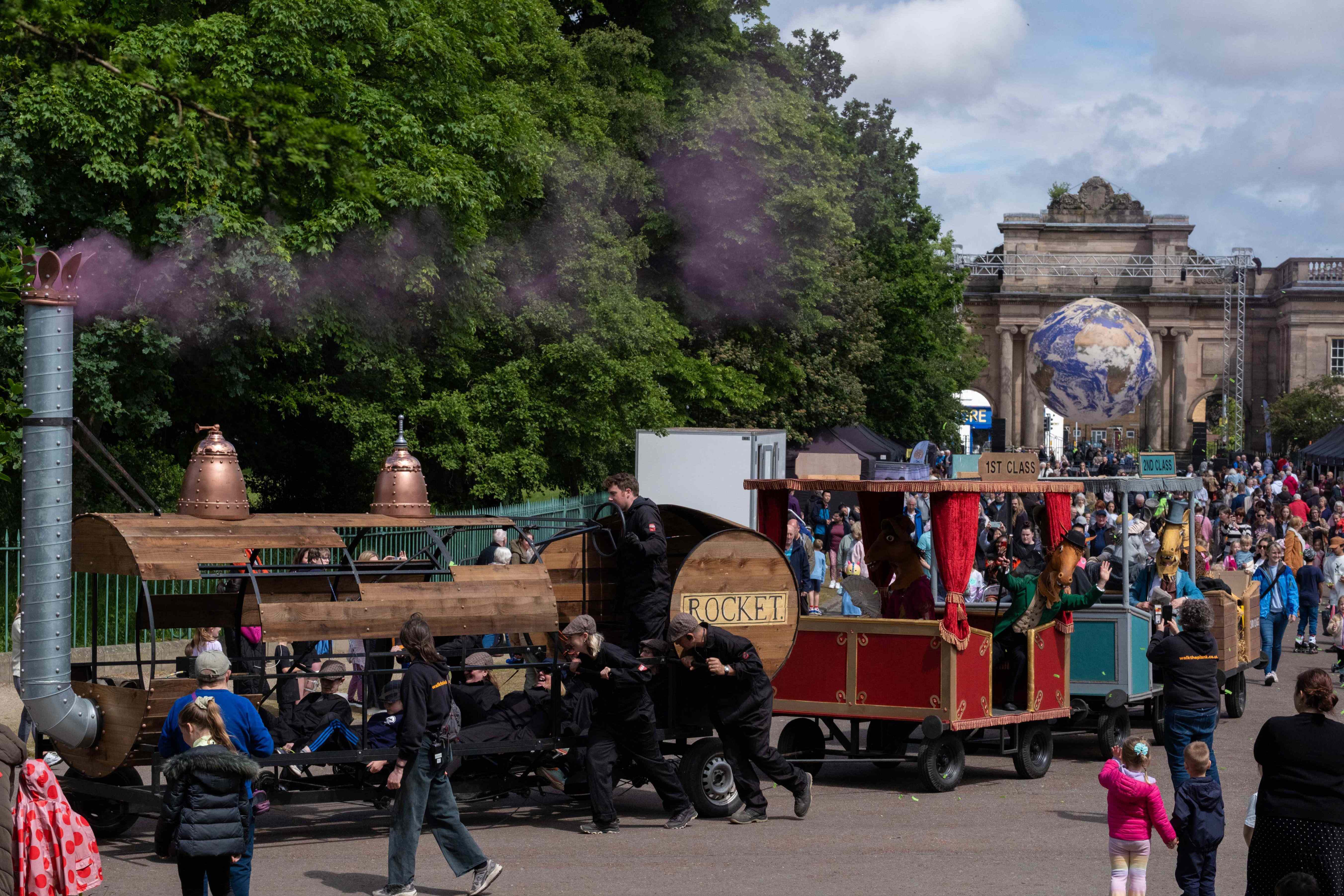 A train float with three carriages carrying pantomime horses in park as crowd look on.