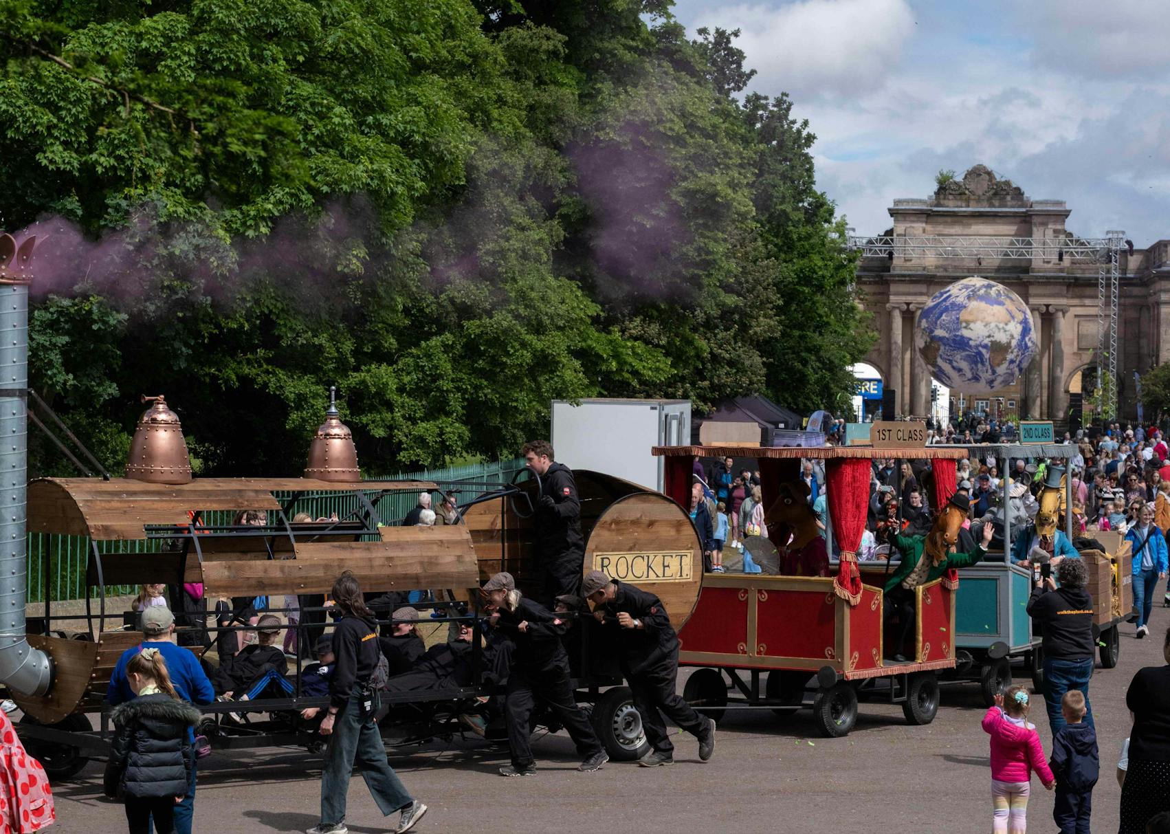 A train float with three carriages carrying pantomime horses in park as crowd look on.