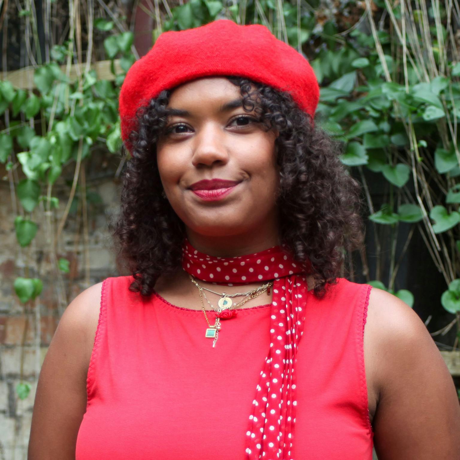 A person standing in front of greenery, she is wearing a red top, a red spotted, scarf and a red beret. She is smiling with her hands in her pockets