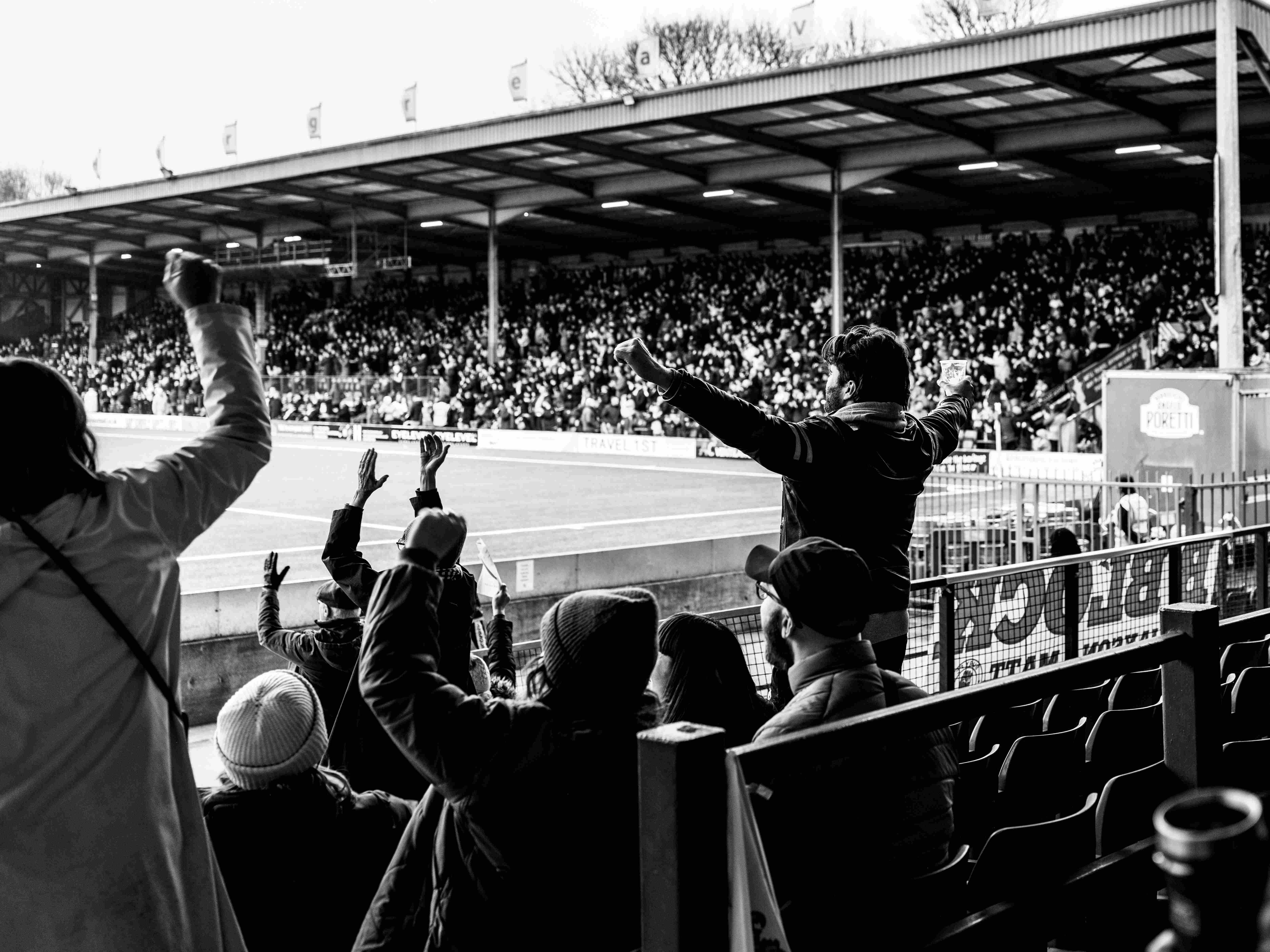 People cheering from the stands of a football pitch with their hands in the air. 