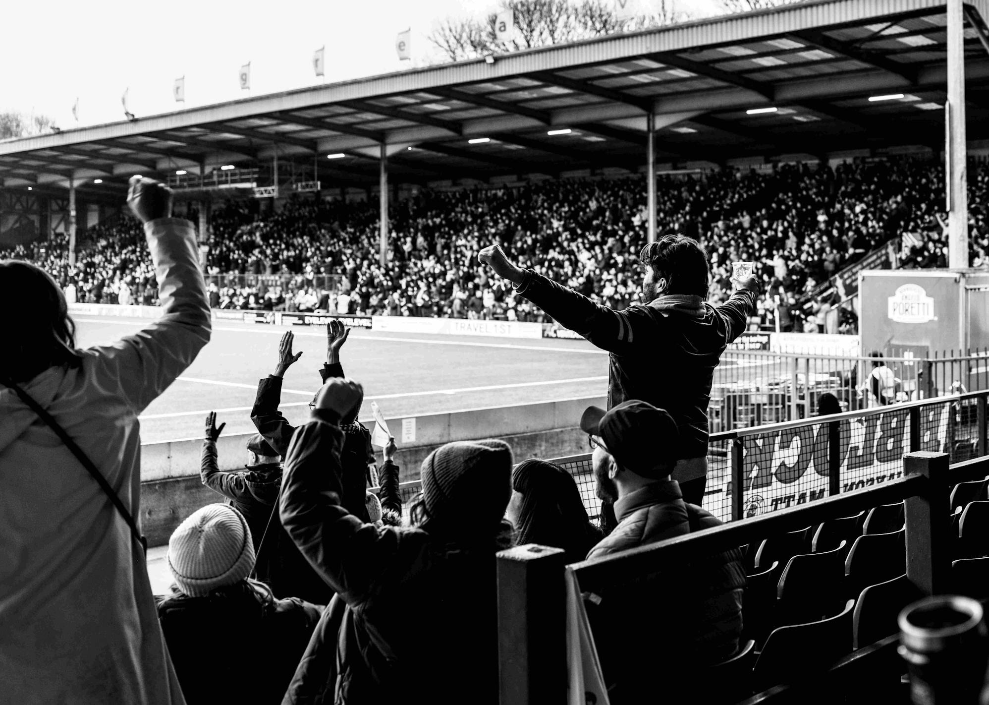 People cheering from the stands of a football pitch with their hands in the air.