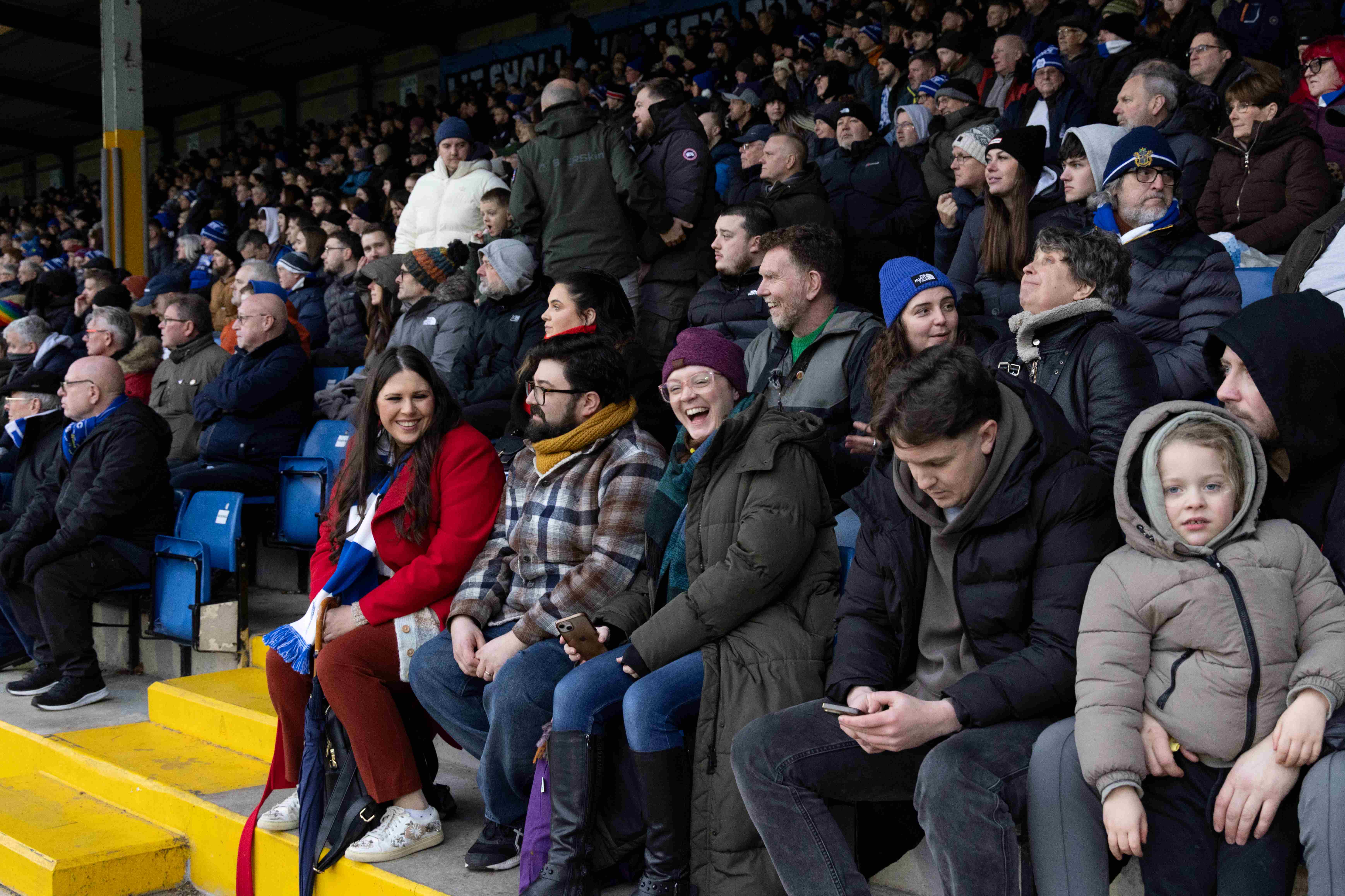 A group of football fans in the stands of a football pitch, smiling and laughing.