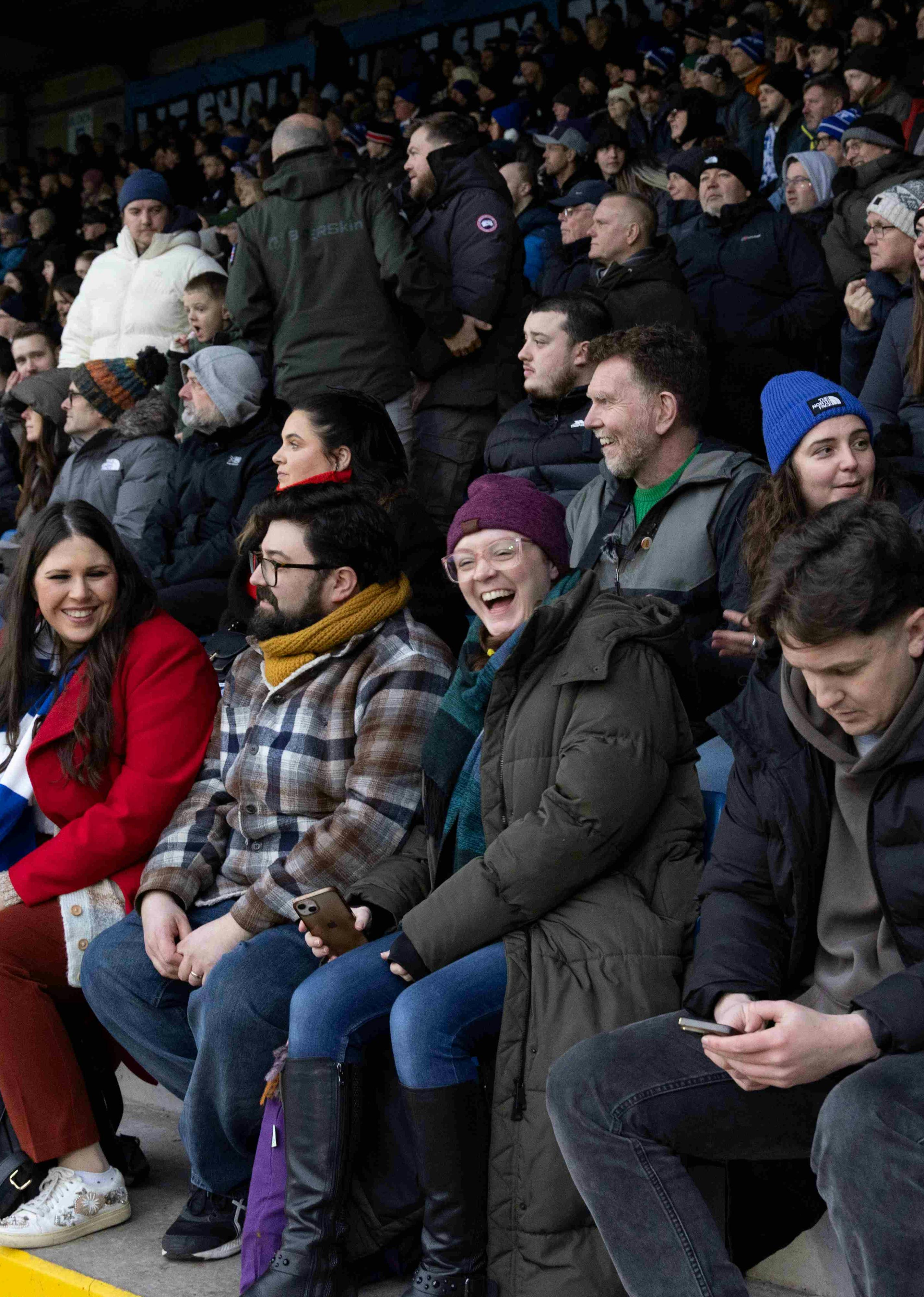A group of football fans in the stands of a football pitch, smiling and laughing.