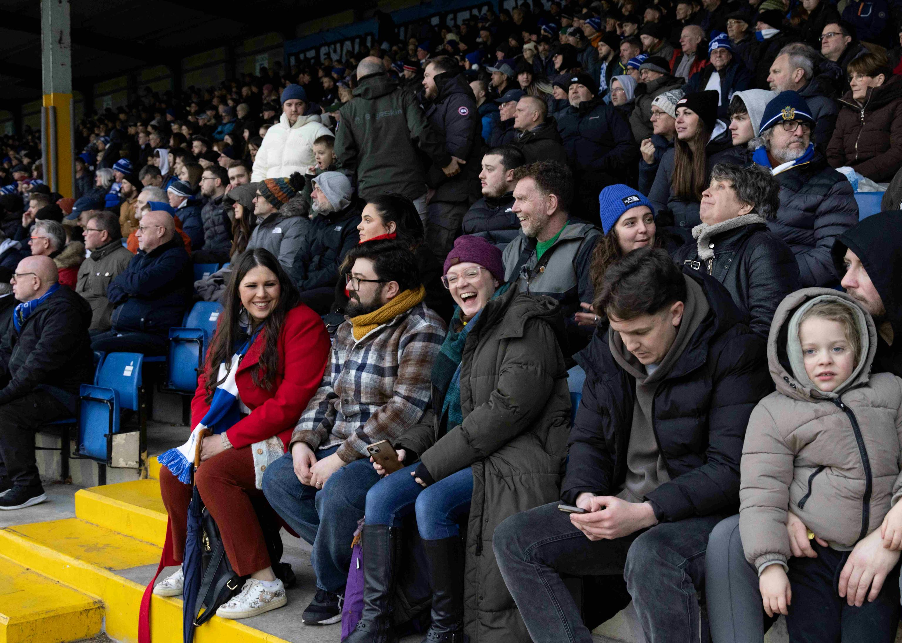 A group of football fans in the stands of a football pitch, smiling and laughing.