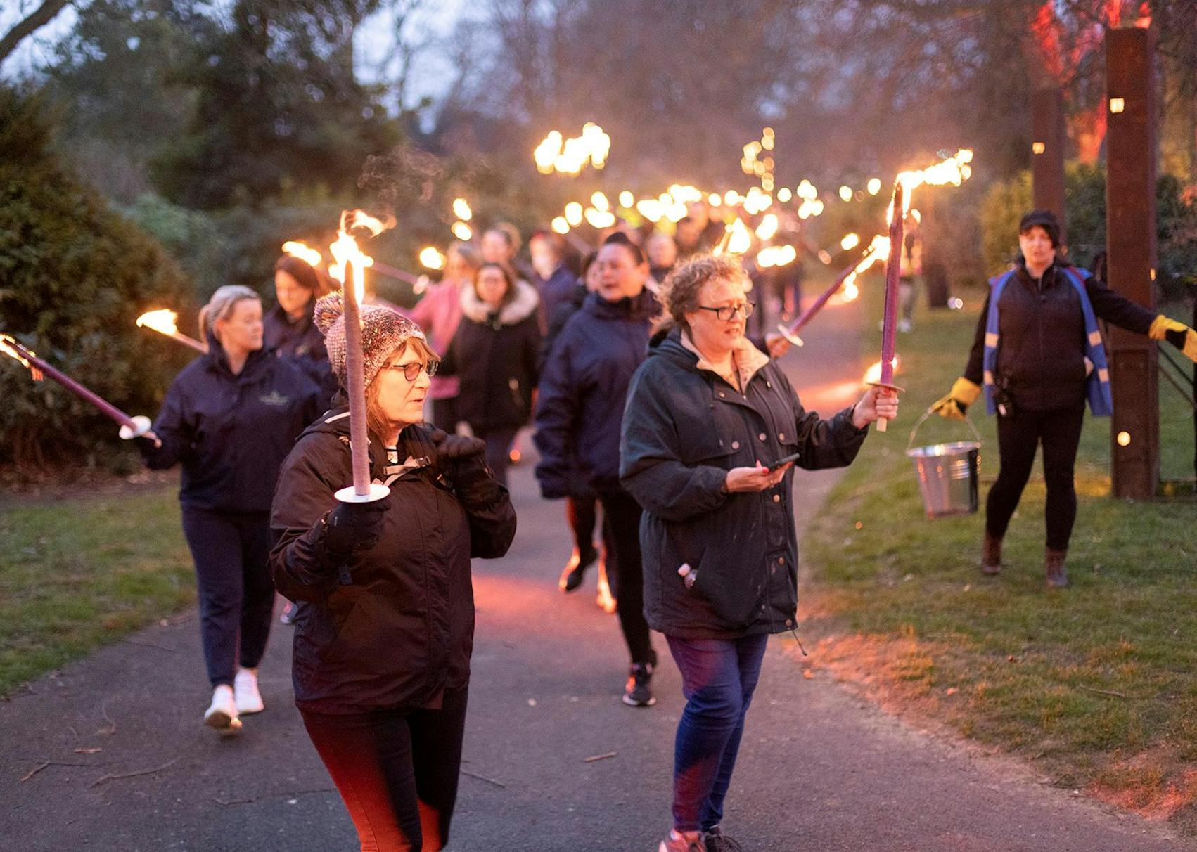 A crowd of people walking in a procession through a park all carrying fire torches