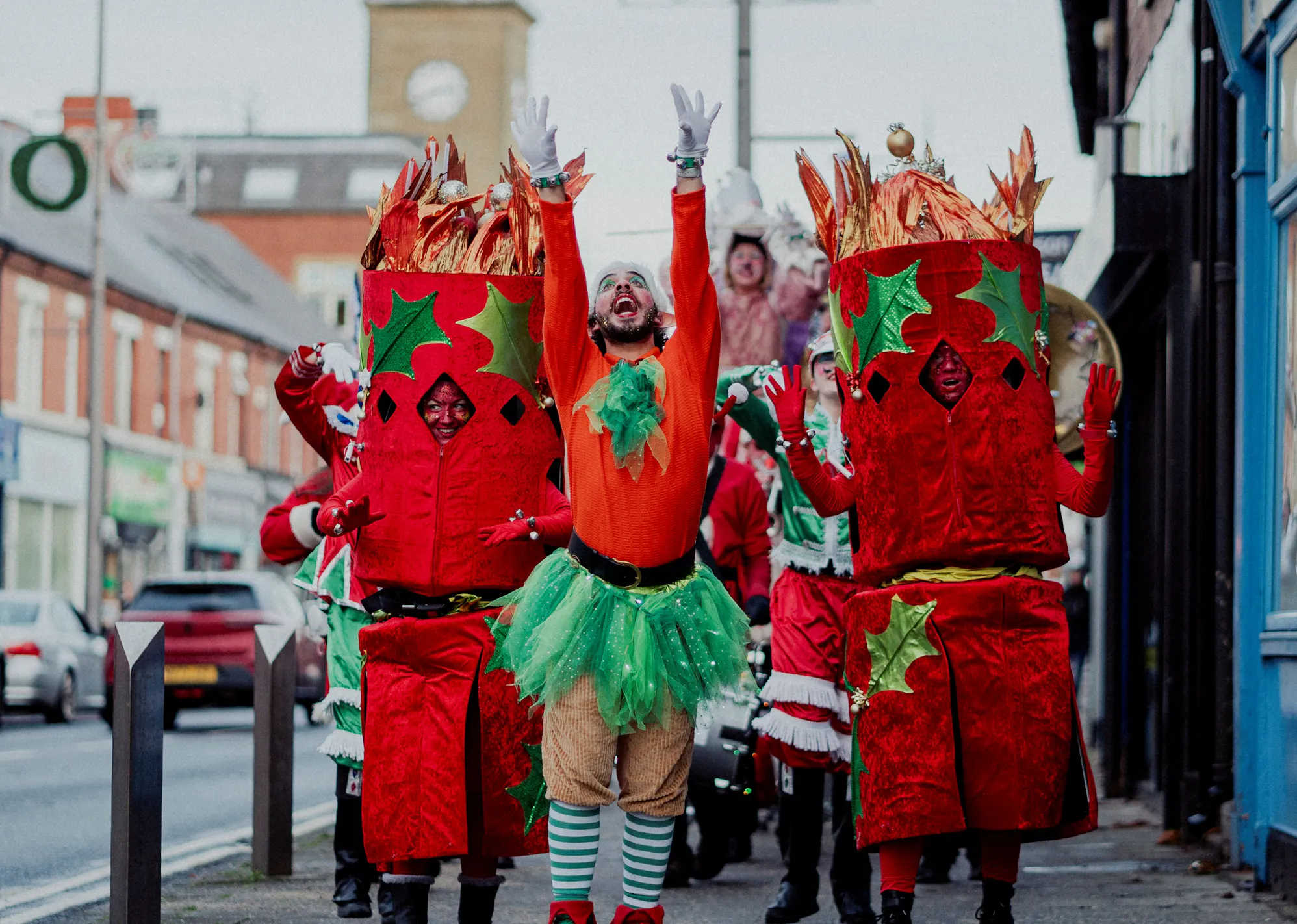 A performer in a red top and green tutu dances down the street flanked by two performers dressed as Christmas Crackers