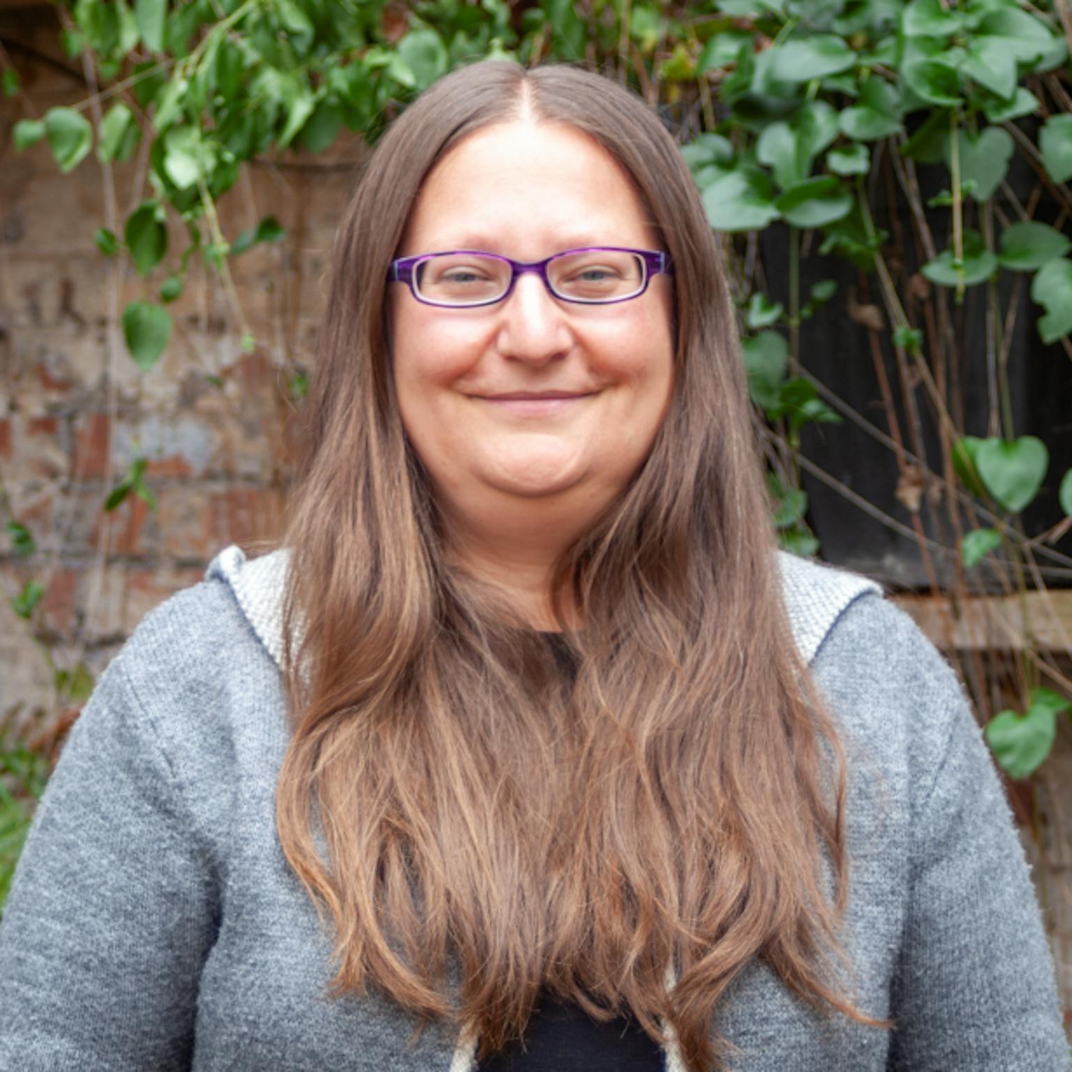 A person with long straight brown hair wearing a grey hoody over a black top standing in front of greenery