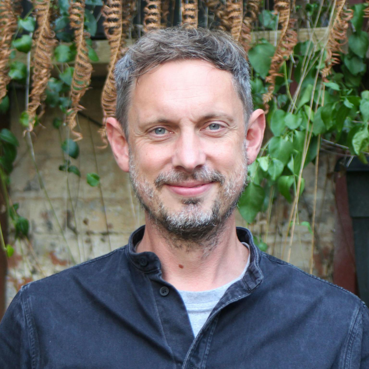 A person with short dark hair and a short beard wearing a dark blue shirt standing in front of greenery