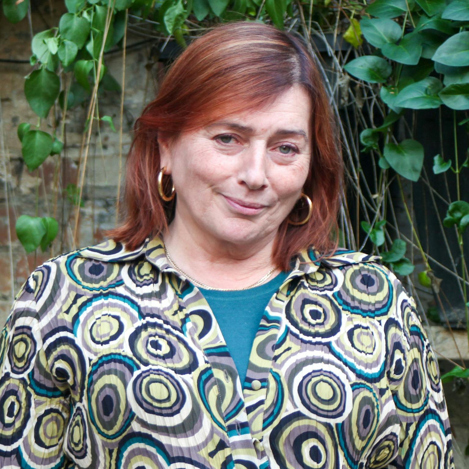 A person standing in front of greenery, she is wearing a green shirt covered in a circular pattern over a blue tee-shirt, she has red hair in a bob. She is smiling with her hands by her sides