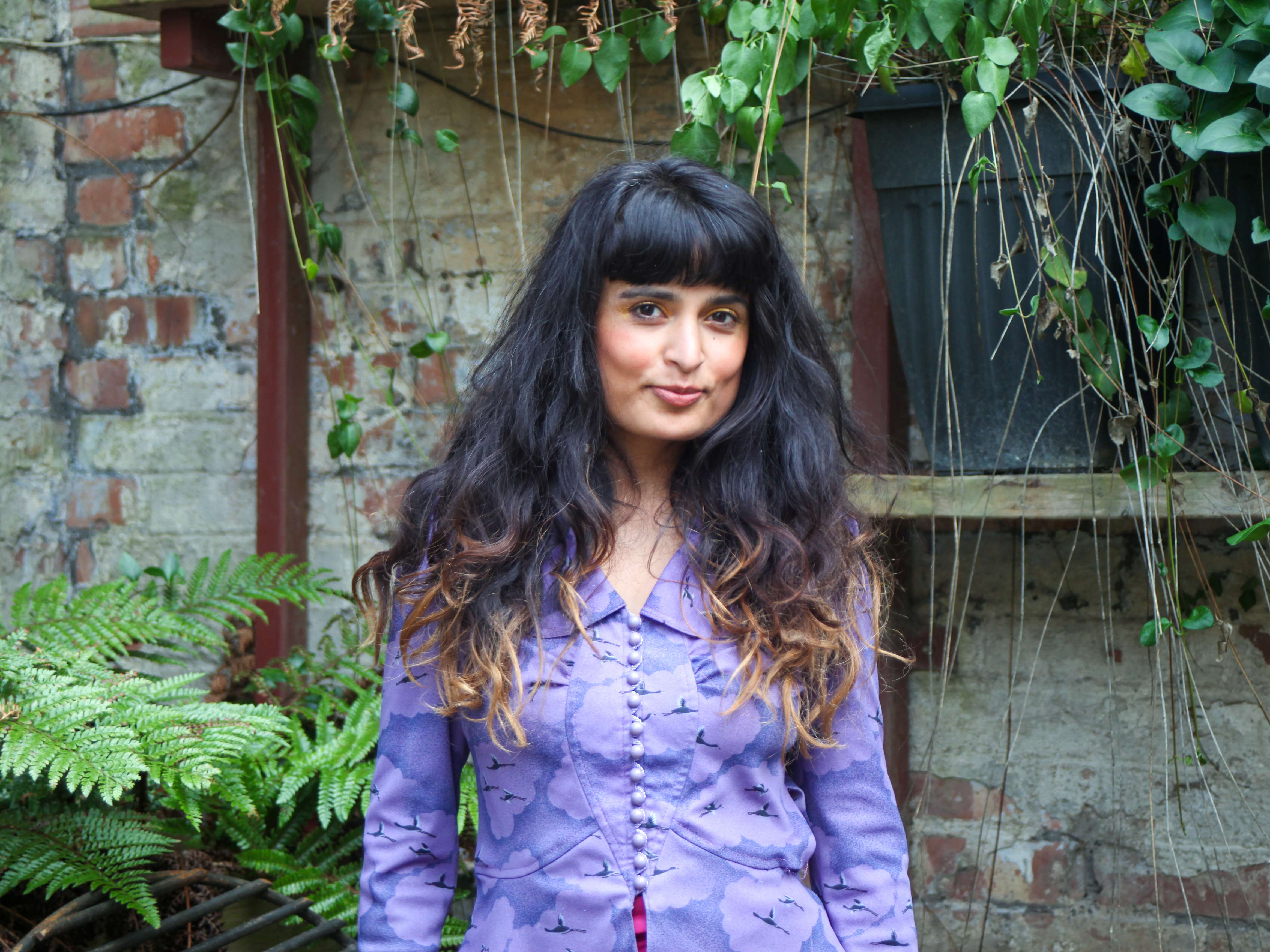 A person standing in front of greenery, with long black hair with blonde at the end, in a purple shirt.