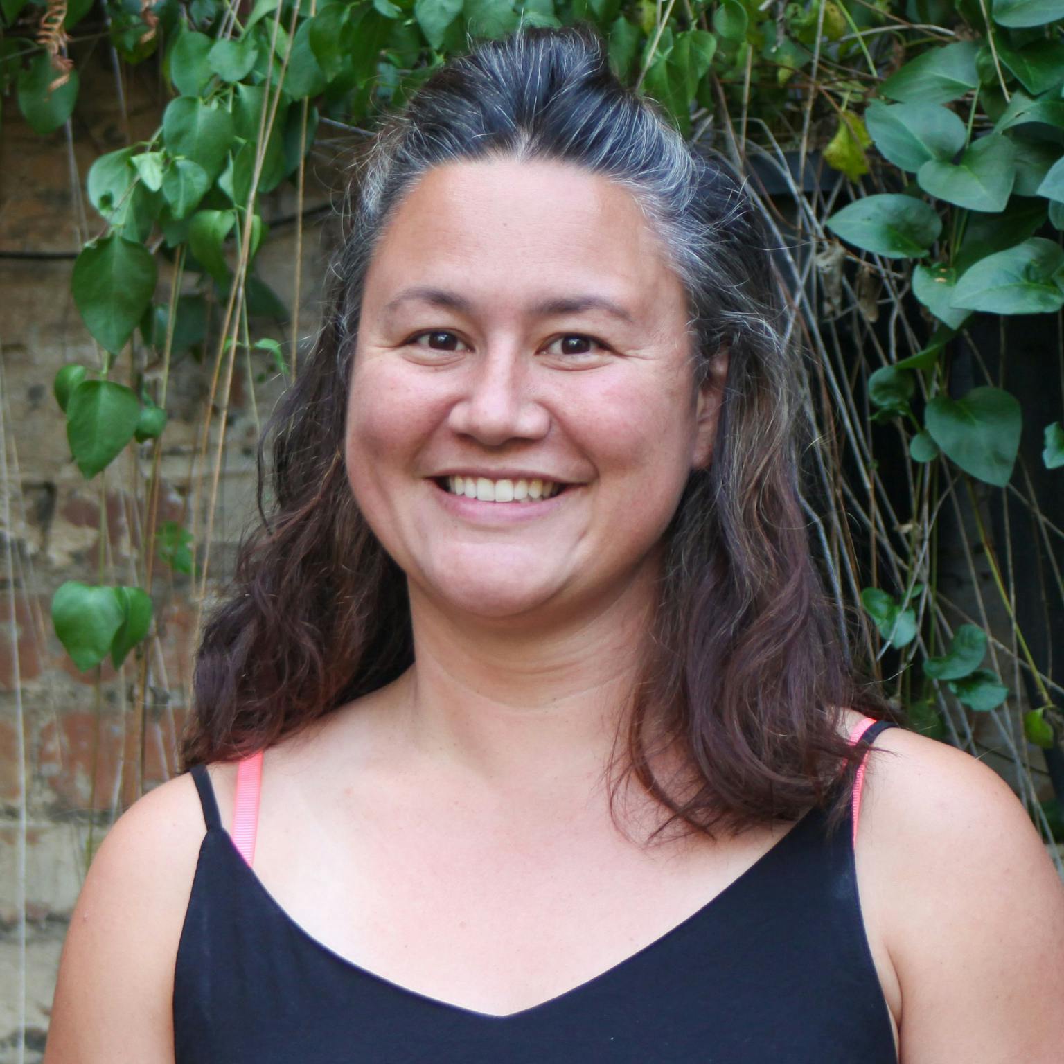 A person standing in front of greenery, she is wearing a black vest top and has dark brown hair that is half pulled-up. She is smiling with her hands by her sides