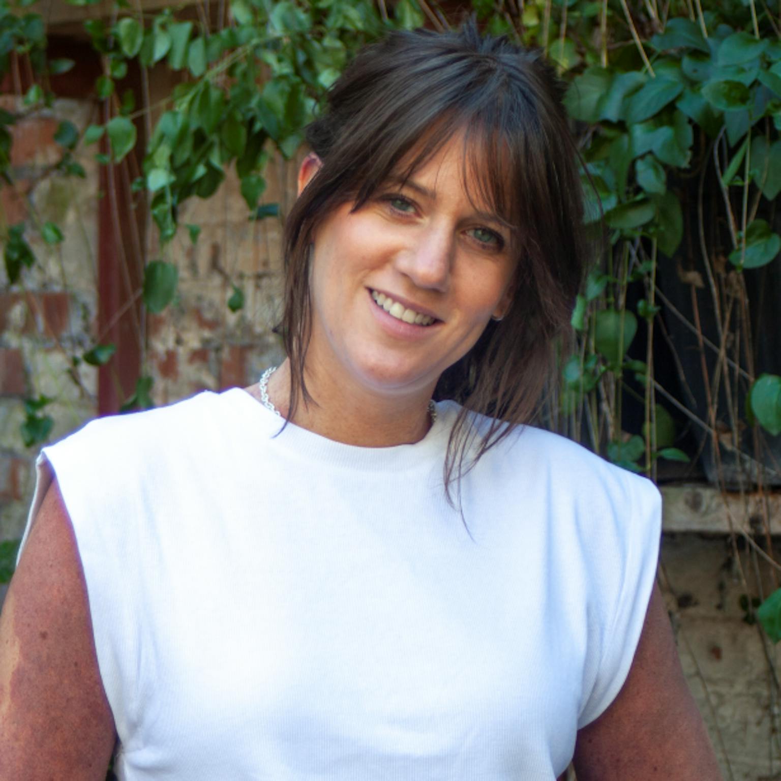A person standing in front of greenery, she is wearing a white sleeveless tee-shirt and has dark brown hair that is up in a bun. She is smiling with her hands behind her back