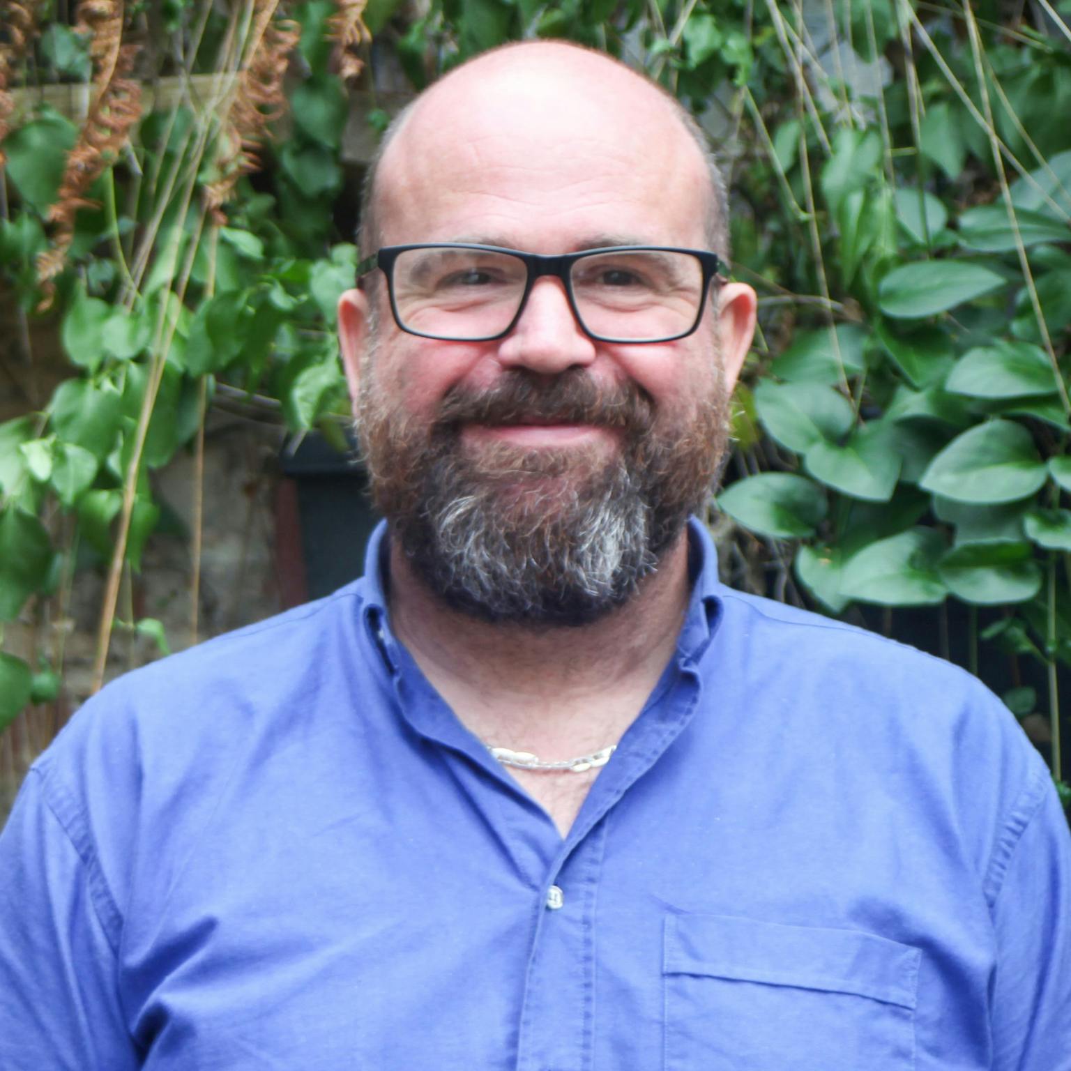 A person standing in front of greenery, he is wearing a blue shirt and a pair of square glasses, he has a dark brown beard. He is smiling with his hands by his sides