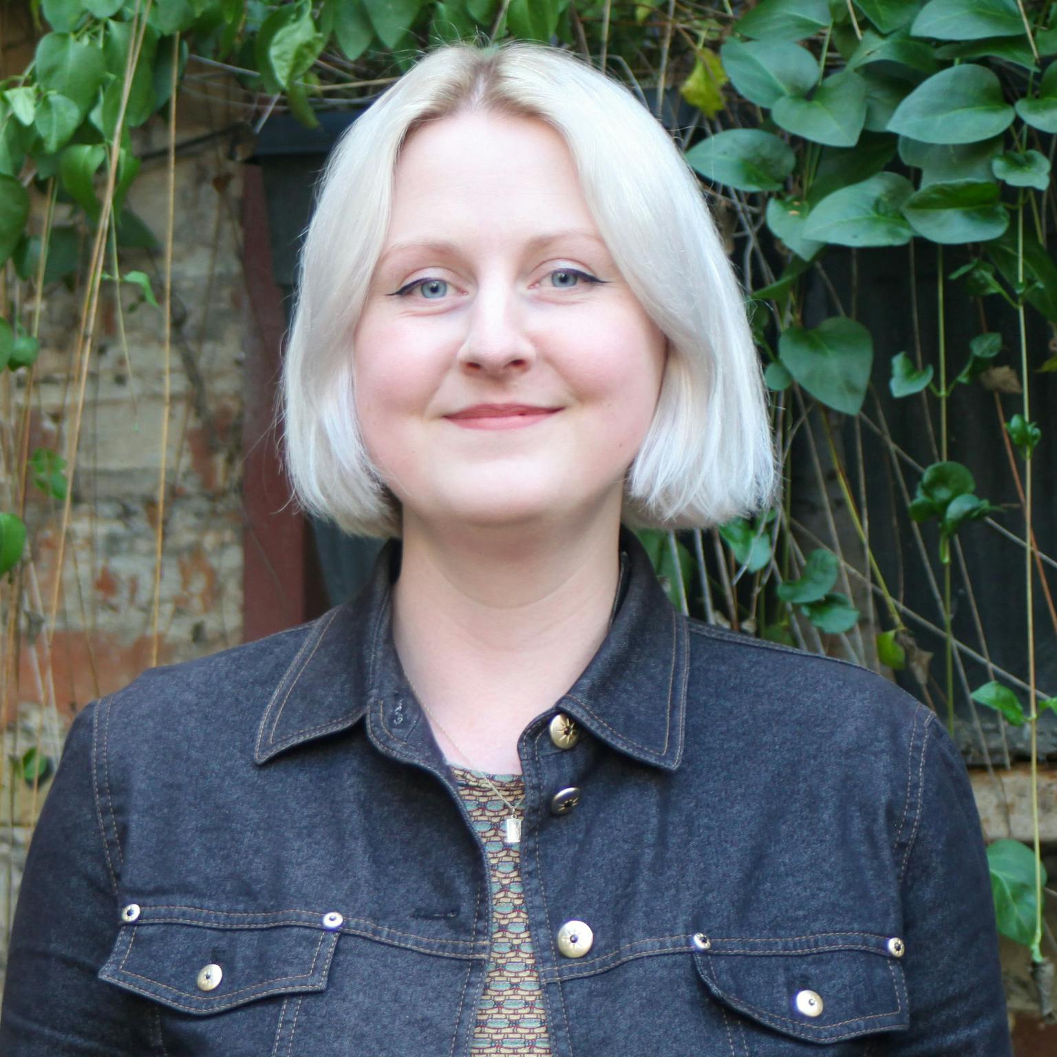 A person standing in front of greenery, she is wearing a dark denim jacket over a patterned top and she has a short platinum-blonde bob. She is smiling with her hands by her sides