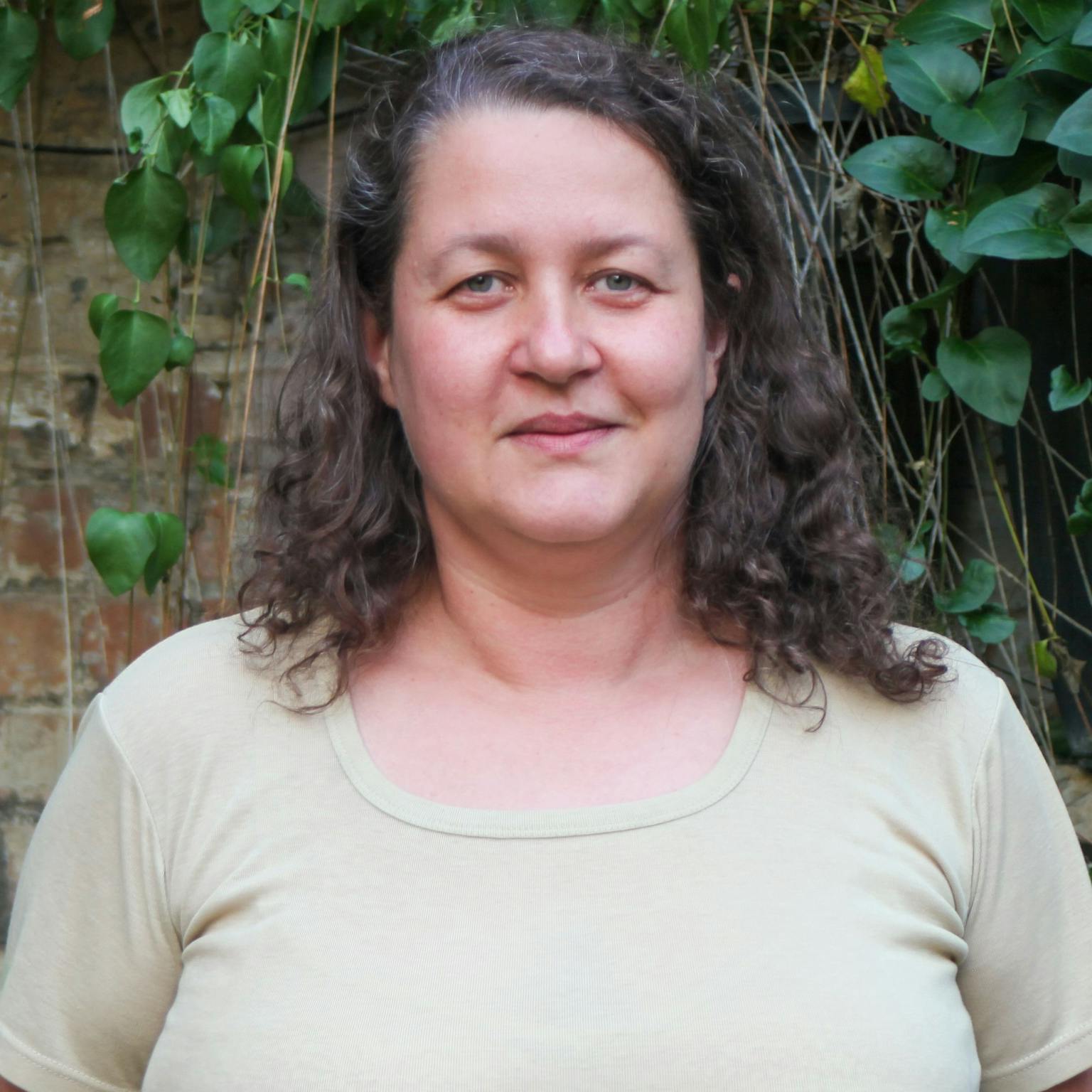 A person standing in front of greenery, she is wearing a light green tee-shirt and has curly brown hair. She is smiling with her hands behind her back