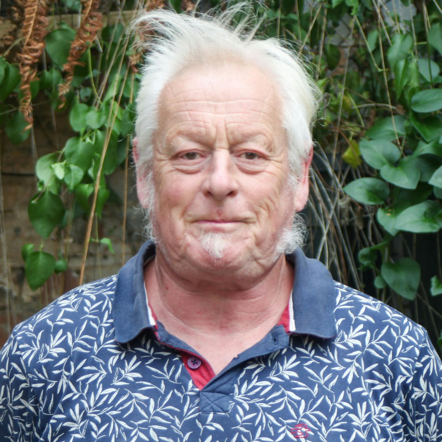 A person standing in front of greenery, he is wearing a patterned blue polo-shirt, he has short white hair and a soul patch. He is smiling with his hands behind his back
