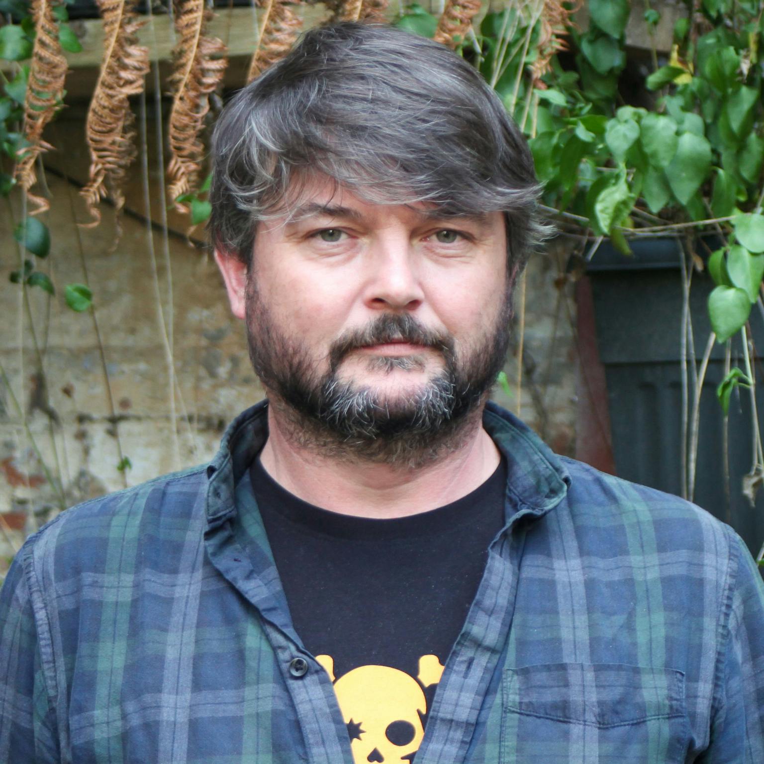 A person standing in front of greenery, he is wearing a dark blue checked shirt over a black tee-shirt with a walk the plank logo on it, he has dark brown hair and a dark brown bear. He is smiling with his arms by his side